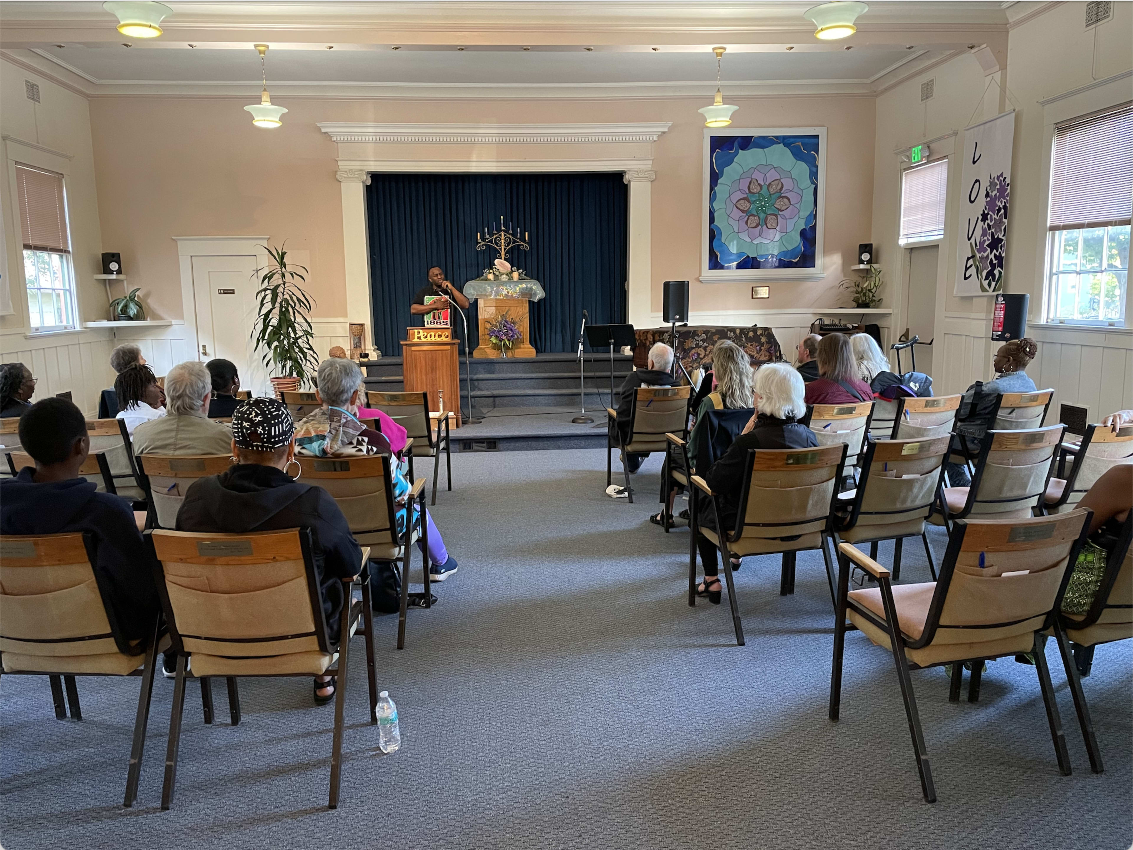 A speaker addresses an audience seated in rows of chairs in a community hall adorned with a floral backdrop and decorative elements.