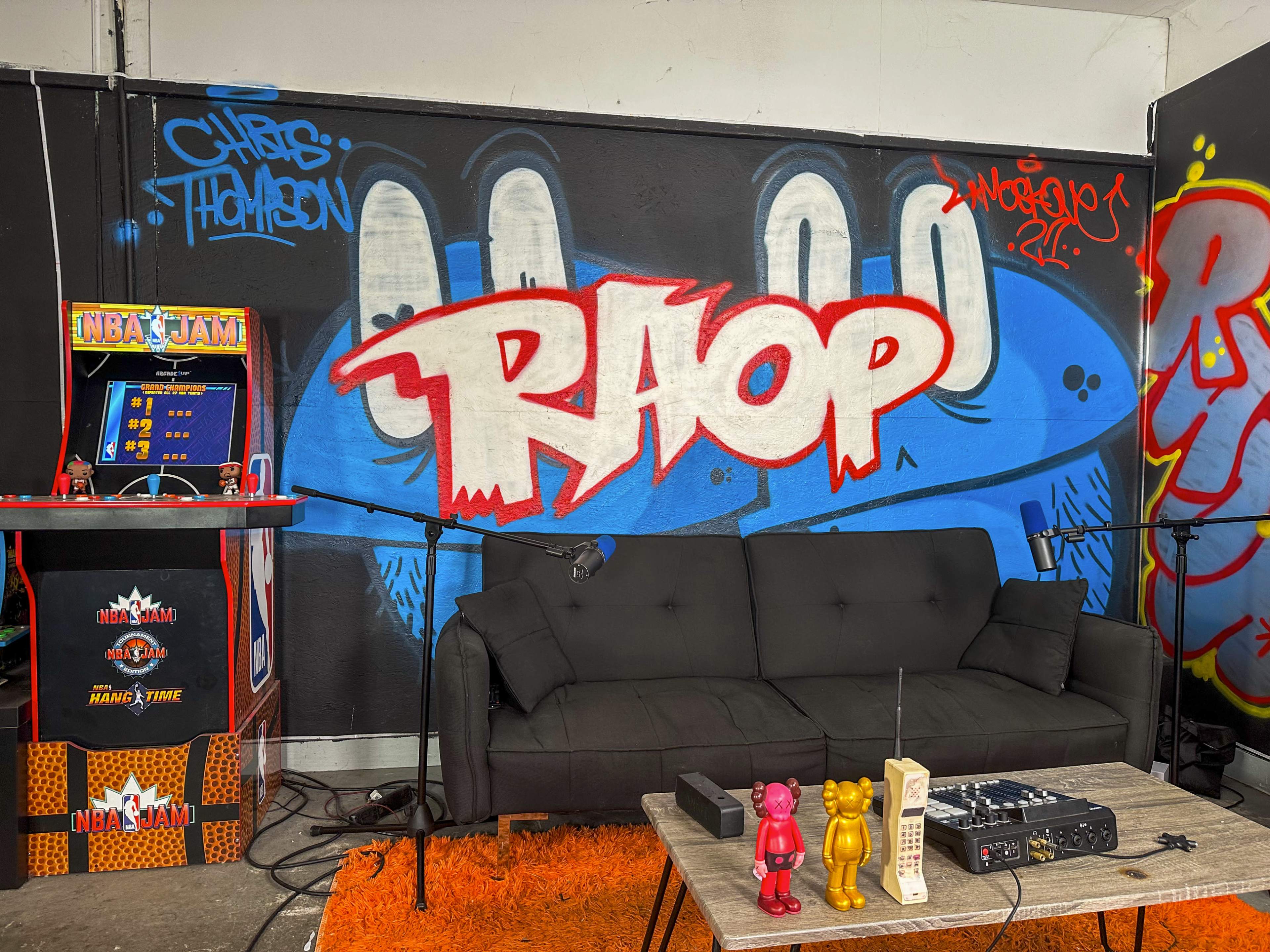 A small studio setup with a black sofa, a microphone, colorful graffiti on the wall, and an NBA Jam arcade game in the corner.