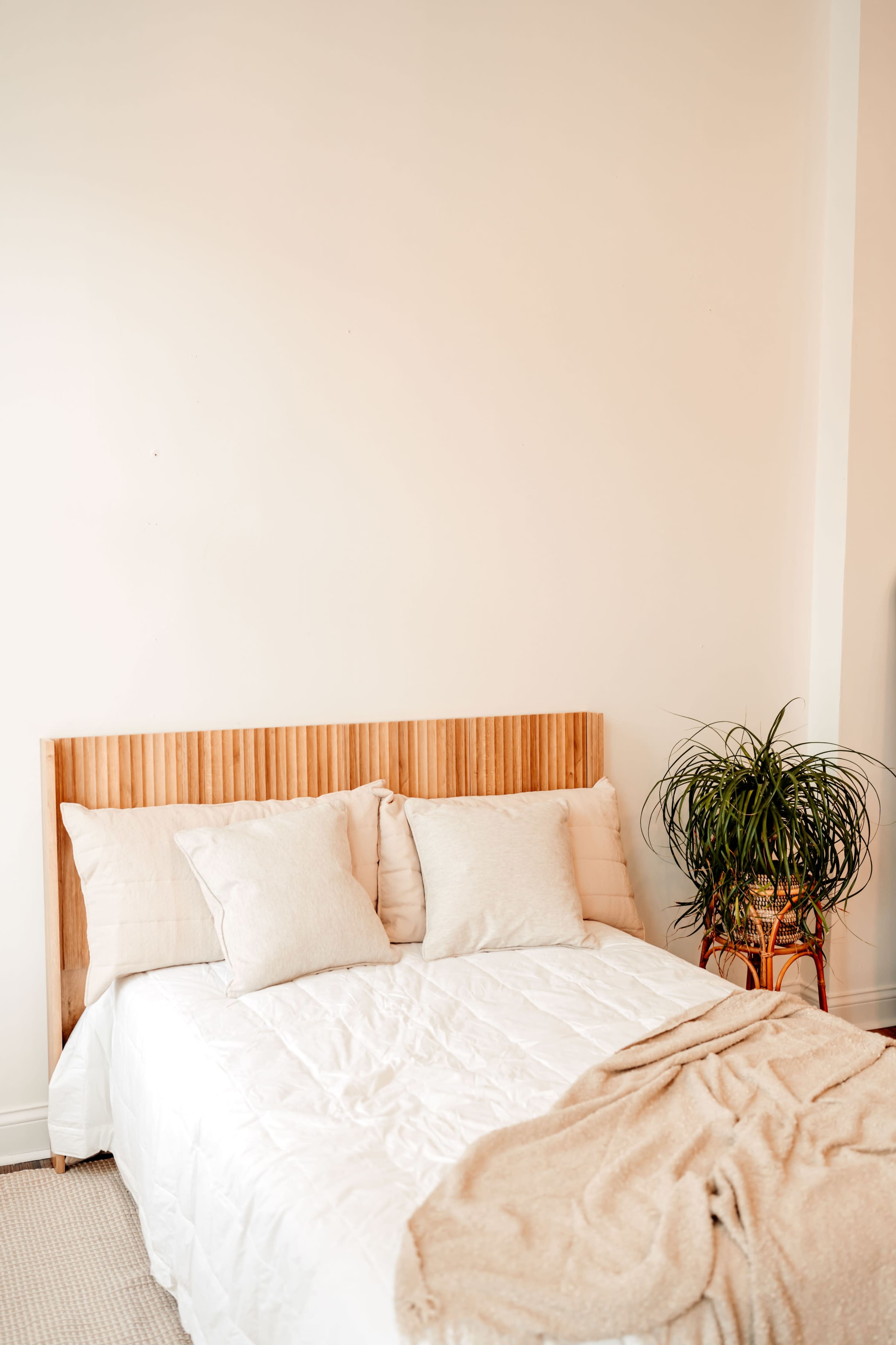 A neatly made bed with beige pillows and a beige throw blanket sits against a light wall, complemented by a potted plant nearby.