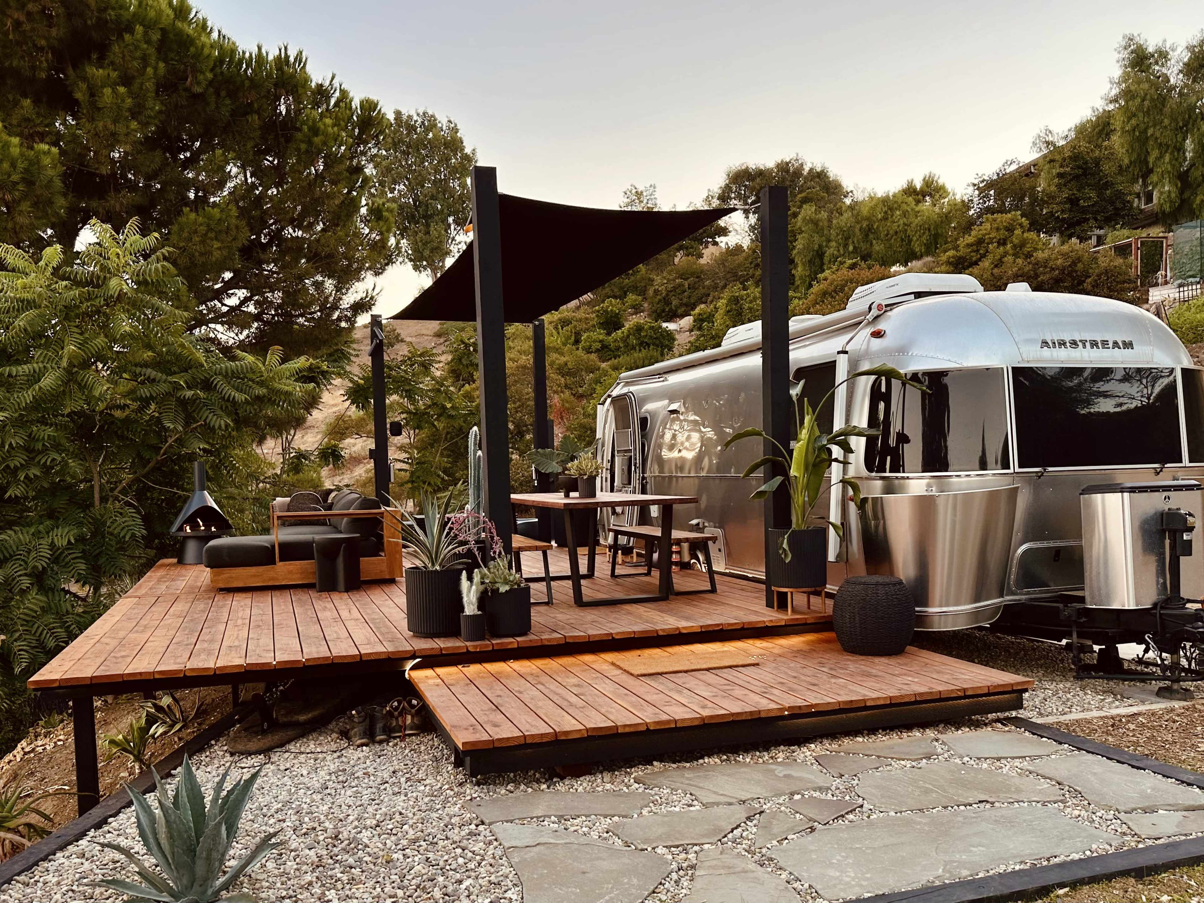 An Airstream trailer is parked next to a wooden deck with outdoor furniture and landscaping, surrounded by trees and stone pathways.