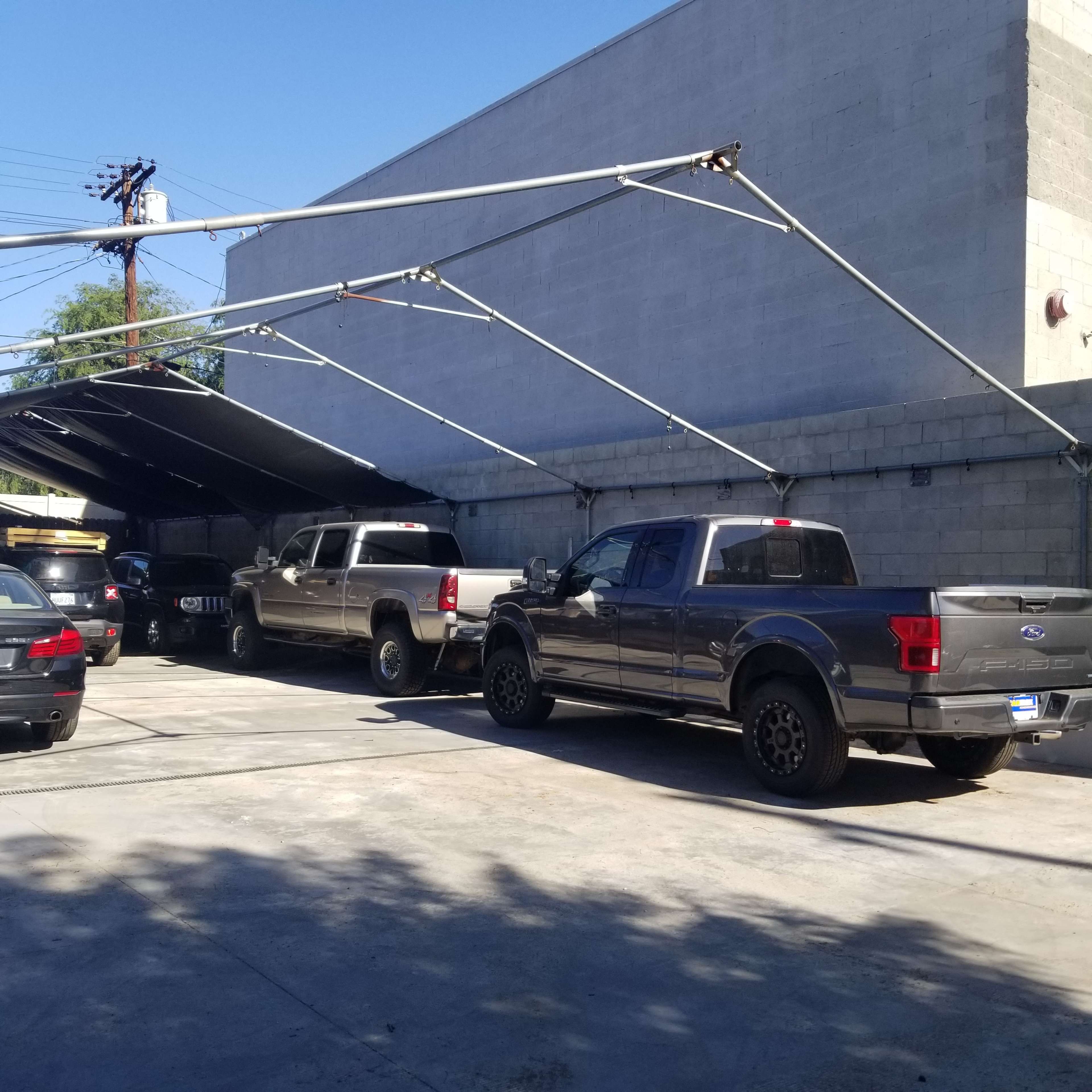 Two pickup trucks are parked under a metal canopy in a concrete lot, with additional vehicles visible in the foreground.