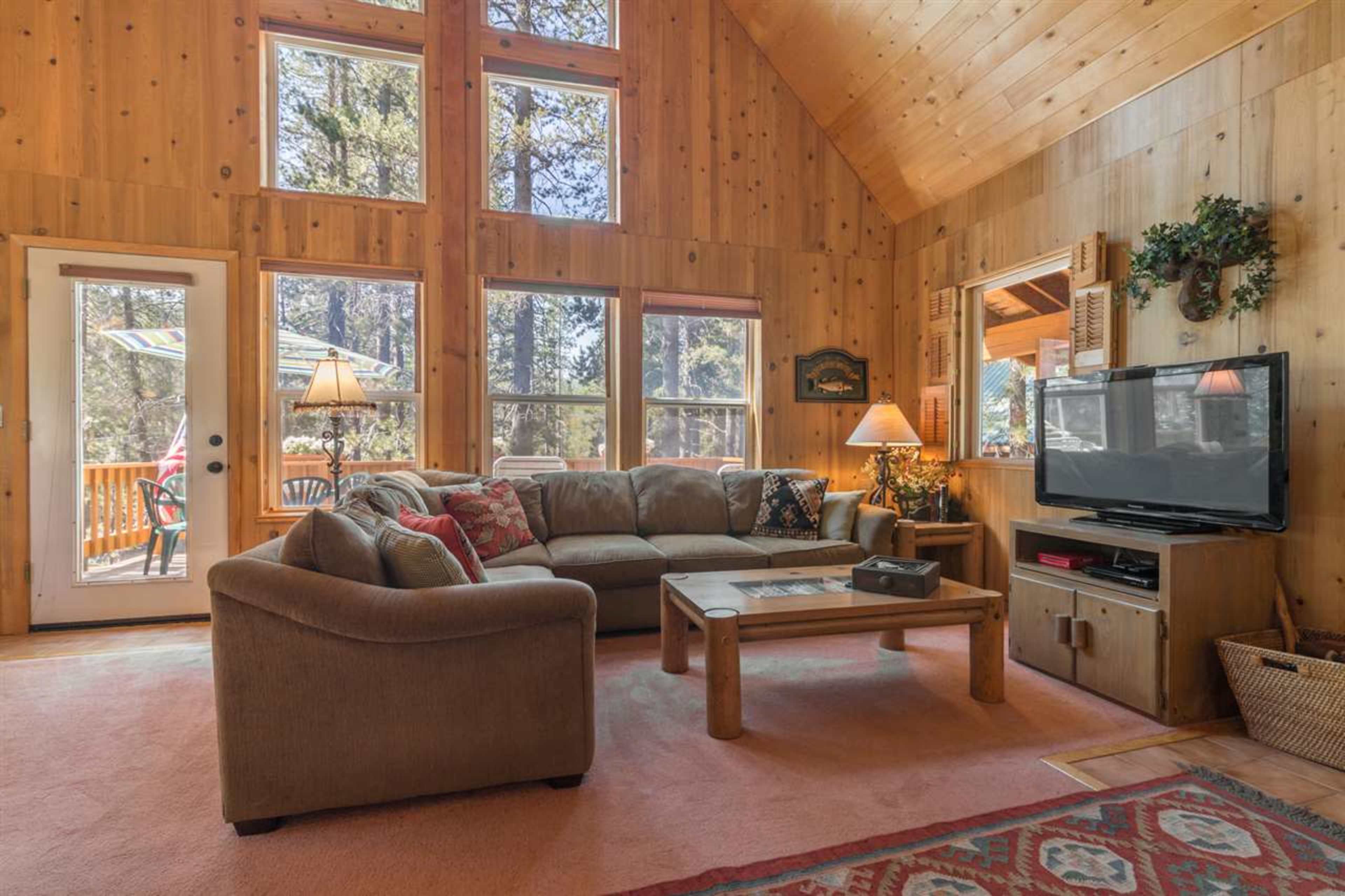 The interior of a wooden cabin features a comfortable living area with a large sofa, a coffee table, and a television, illuminated by natural light from large windows.