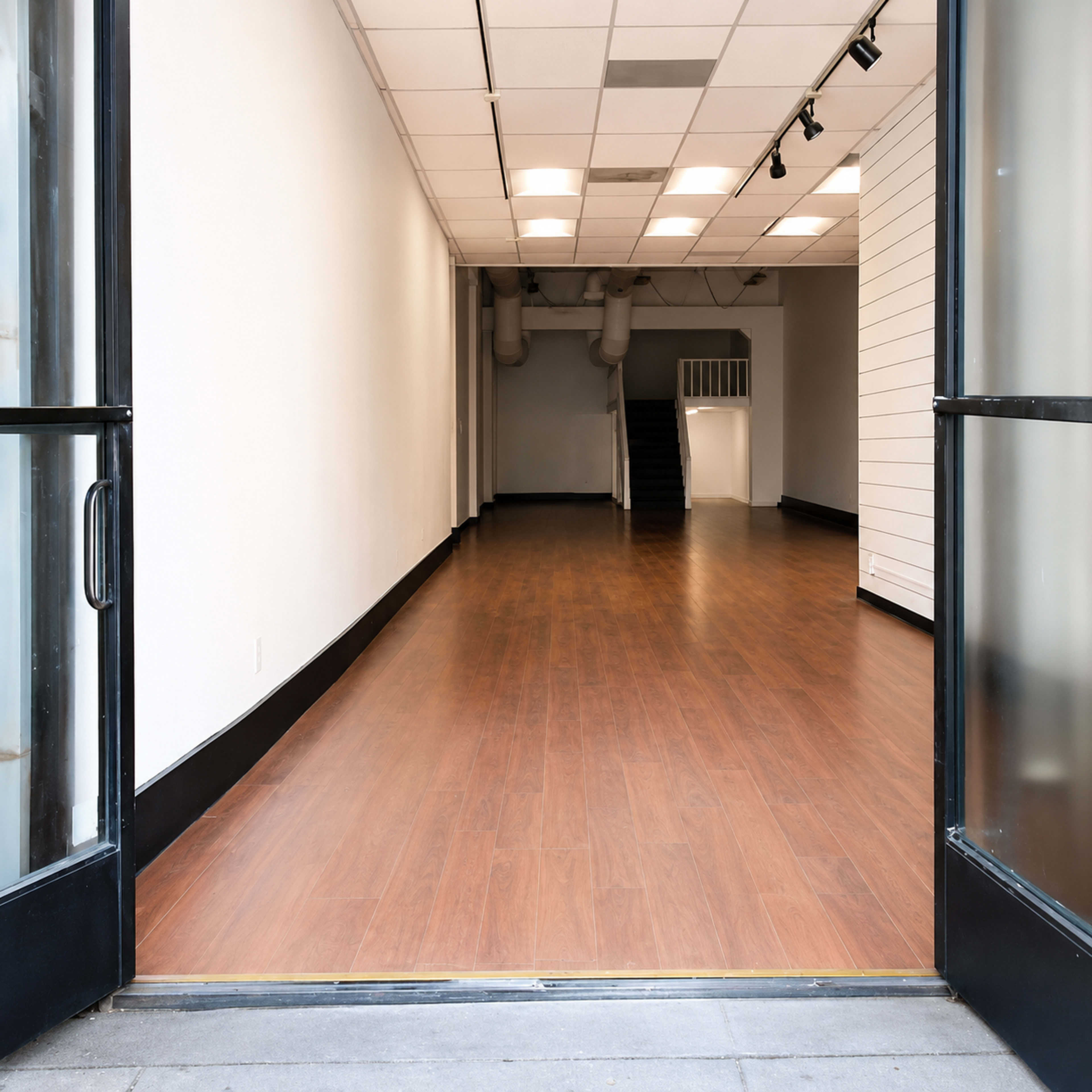 The image shows the interior of an empty retail space with wooden flooring and white walls, viewed through an open glass door.