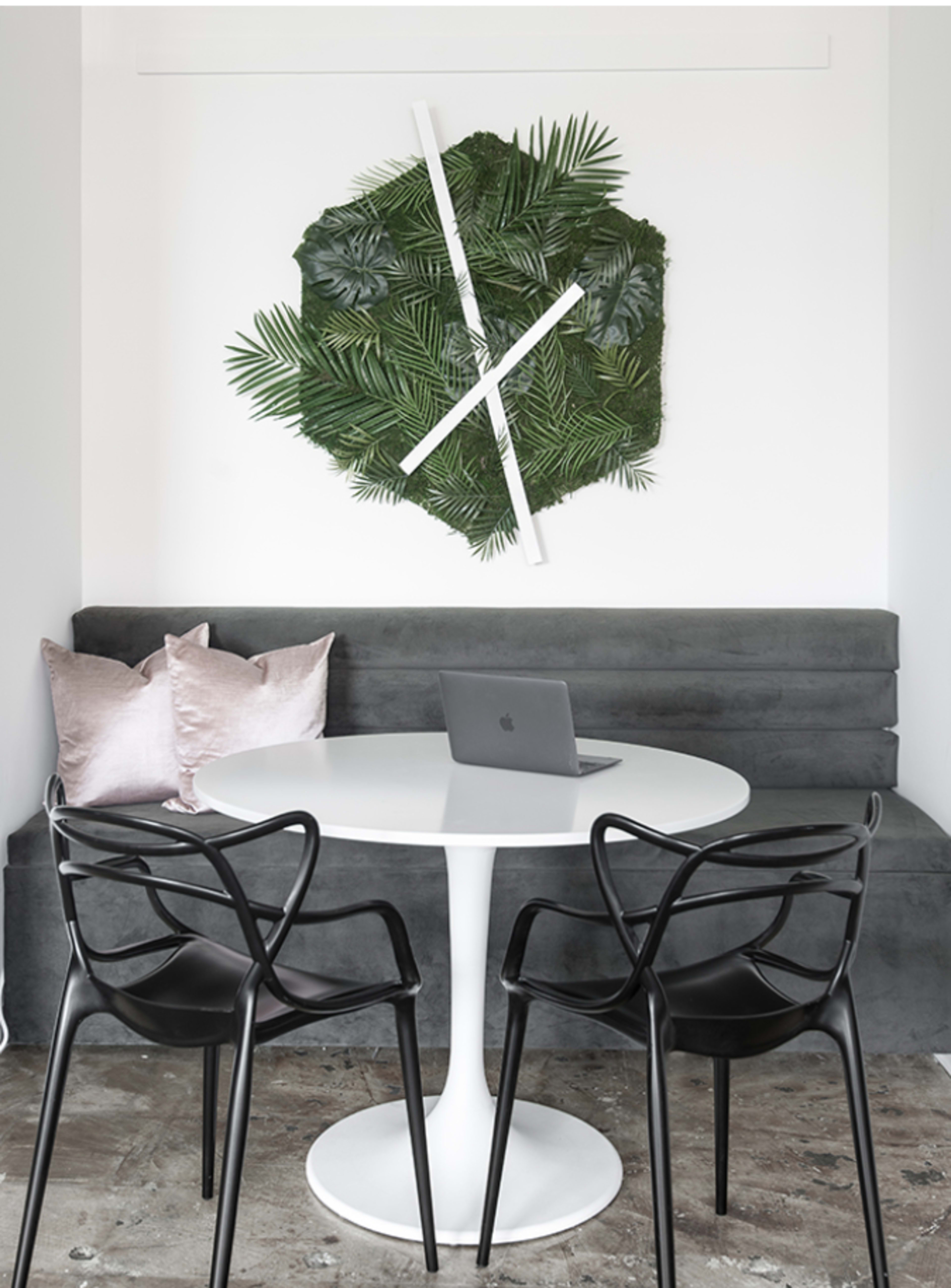 A modern dining area with a round white table, black chairs, a gray bench, a laptop on the table, and a decorative wall arrangement of greenery.