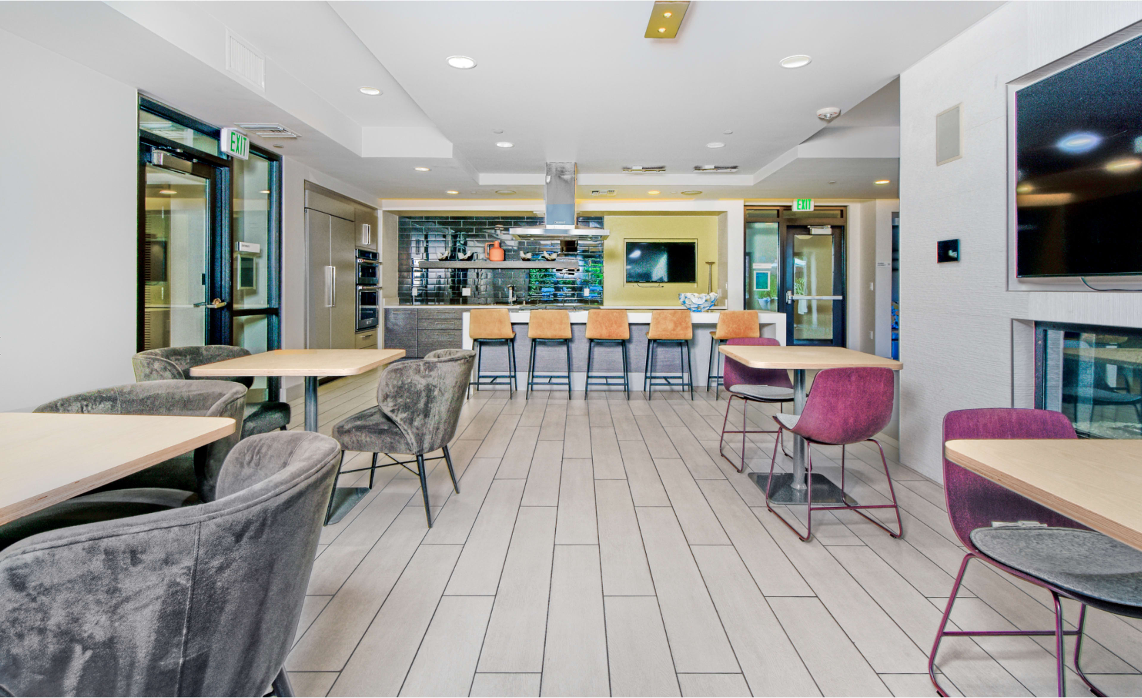 The image features a modern dining area with several tables and chairs arranged around a kitchen area, including bar seating.