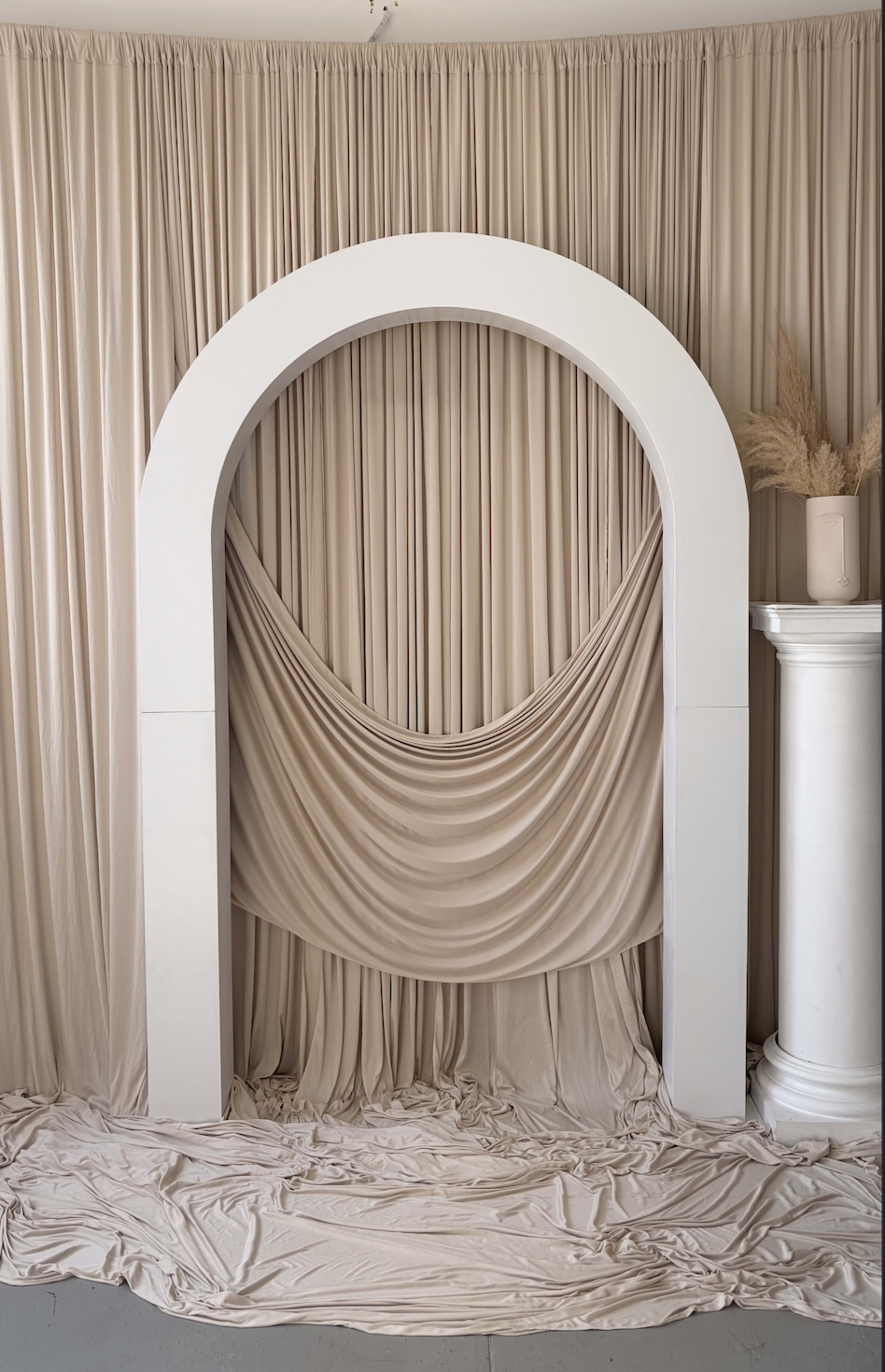 The image features a minimalist arch structure in front of draped fabric and a smooth floor, creating a serene backdrop.