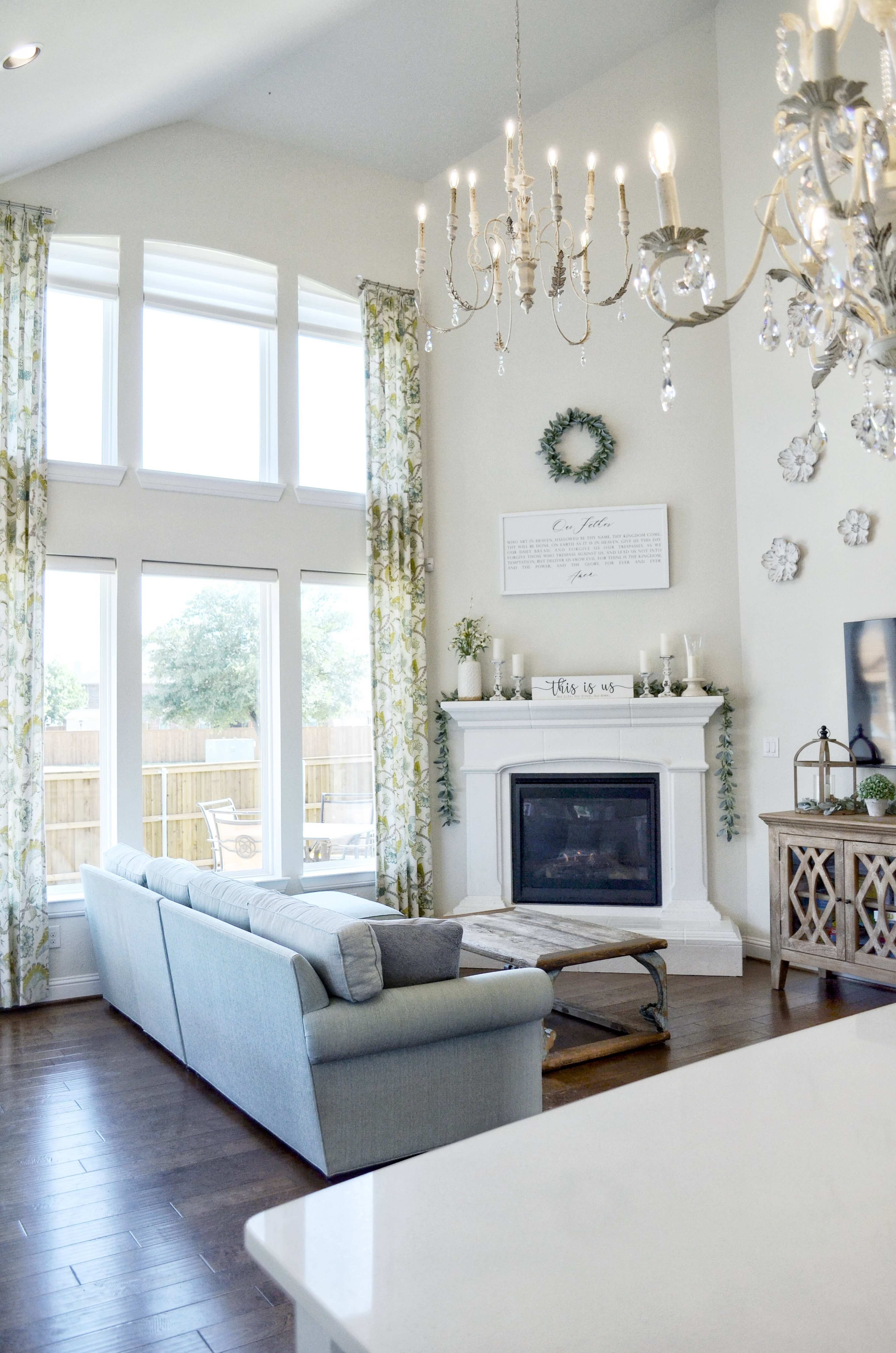 A bright living room features a fireplace surrounded by large windows and decorative curtains, with a light blue sofa and wooden flooring.