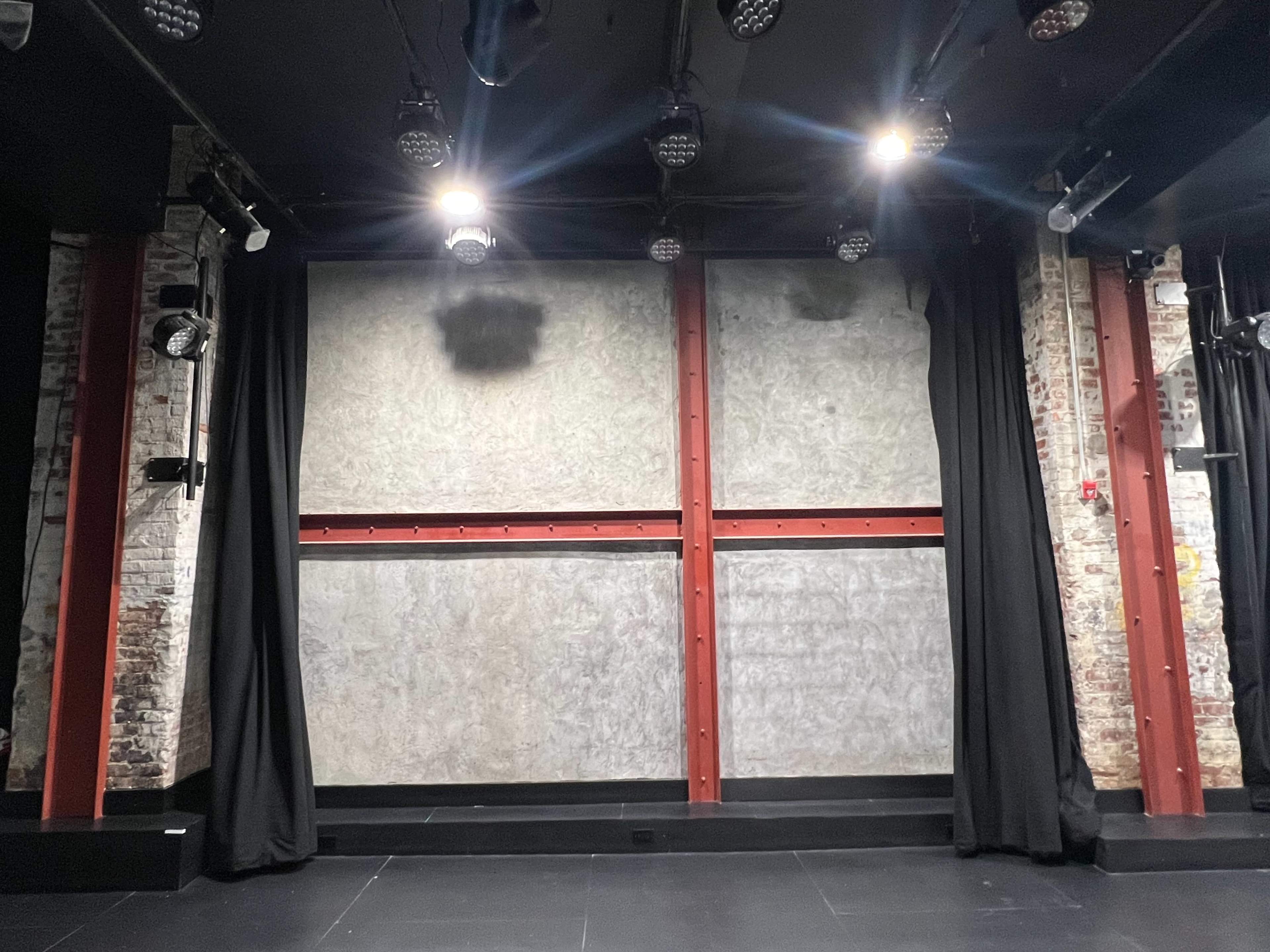 The image shows a theater stage with black curtains and a weathered gray wall, illuminated by overhead stage lights.
