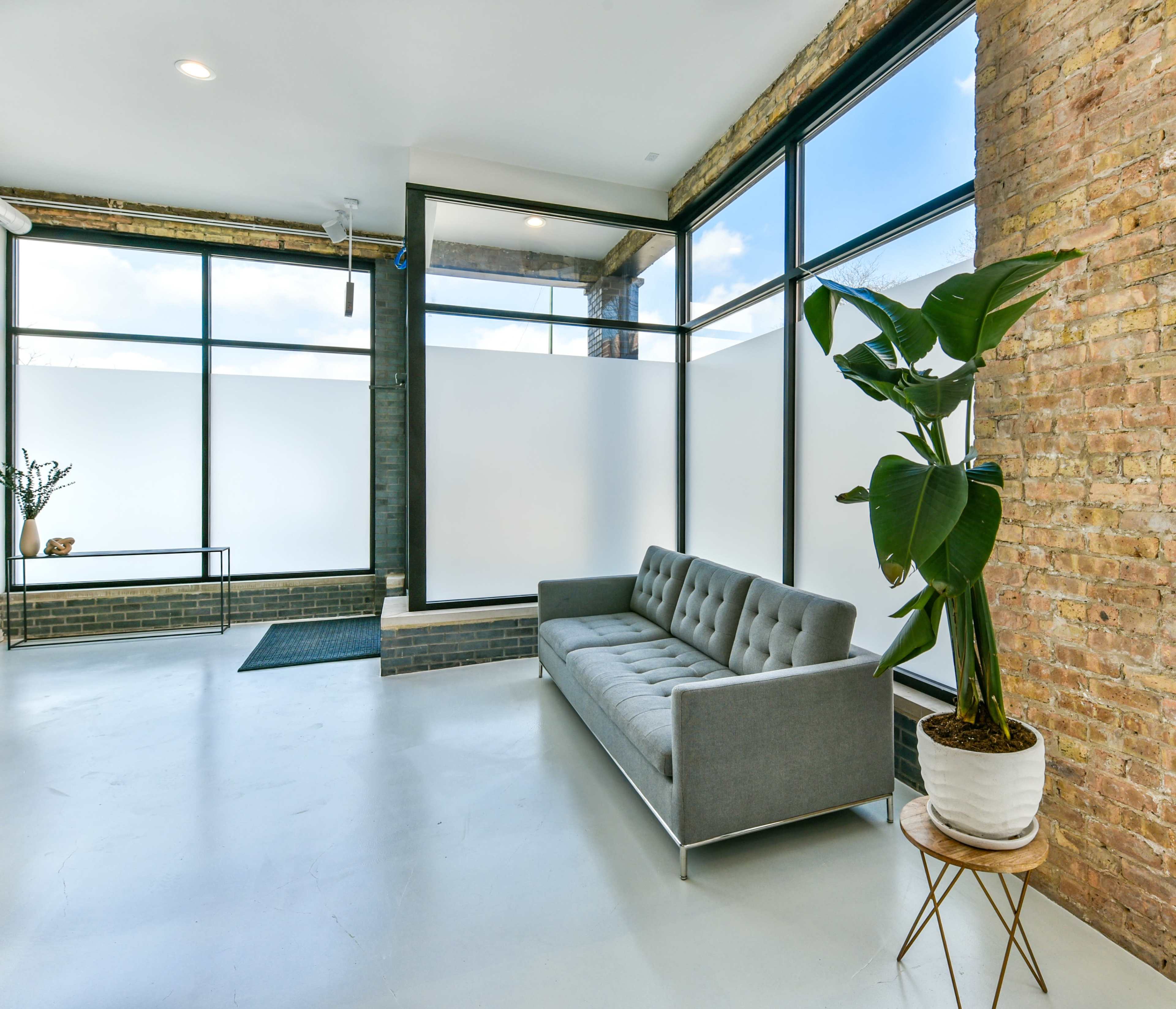 A modern interior space featuring a gray sofa, large windows, a potted plant, and exposed brick walls.