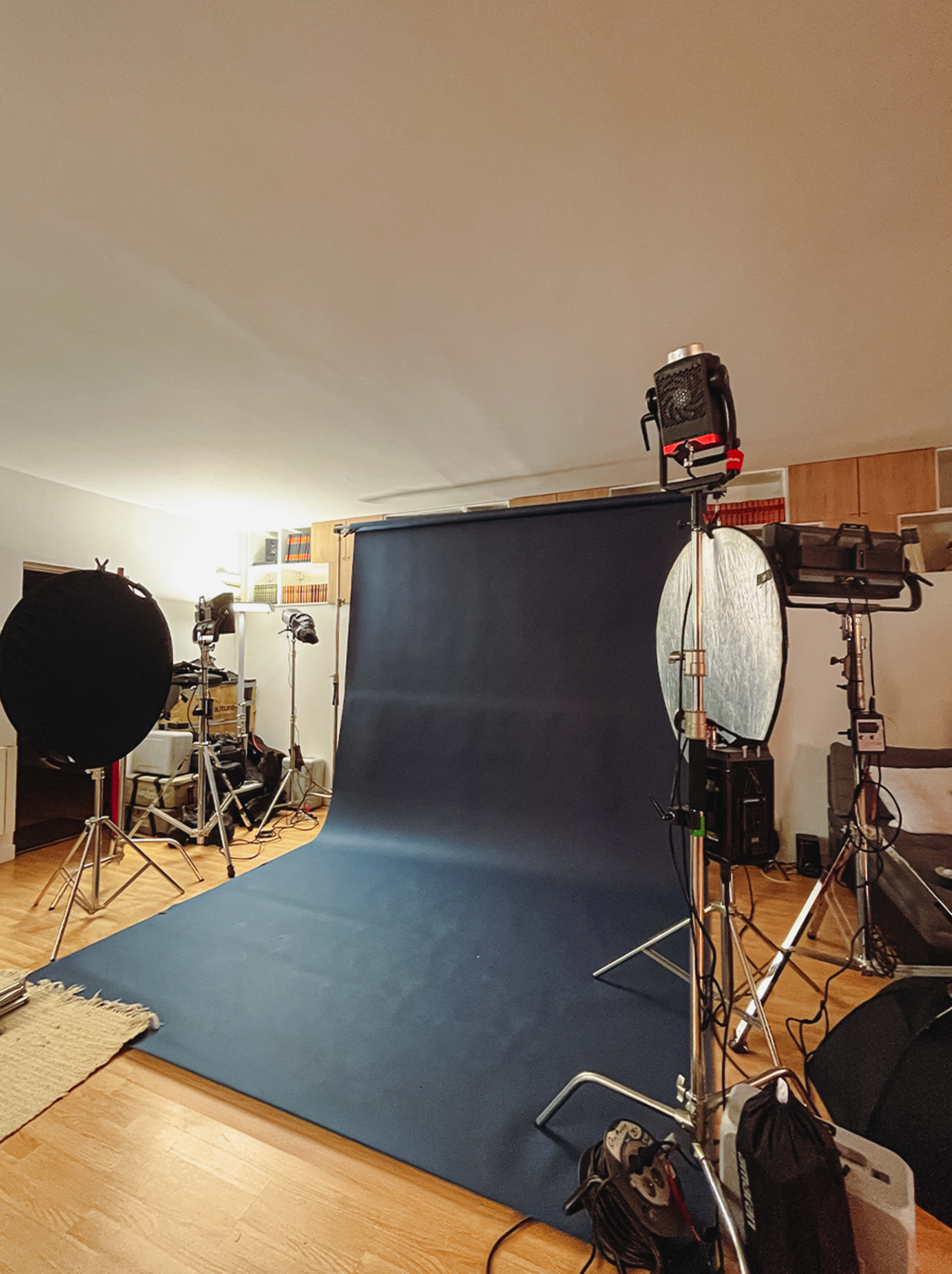The image shows a photography studio setup with a dark backdrop, lighting equipment on stands, and various props arranged in the space.