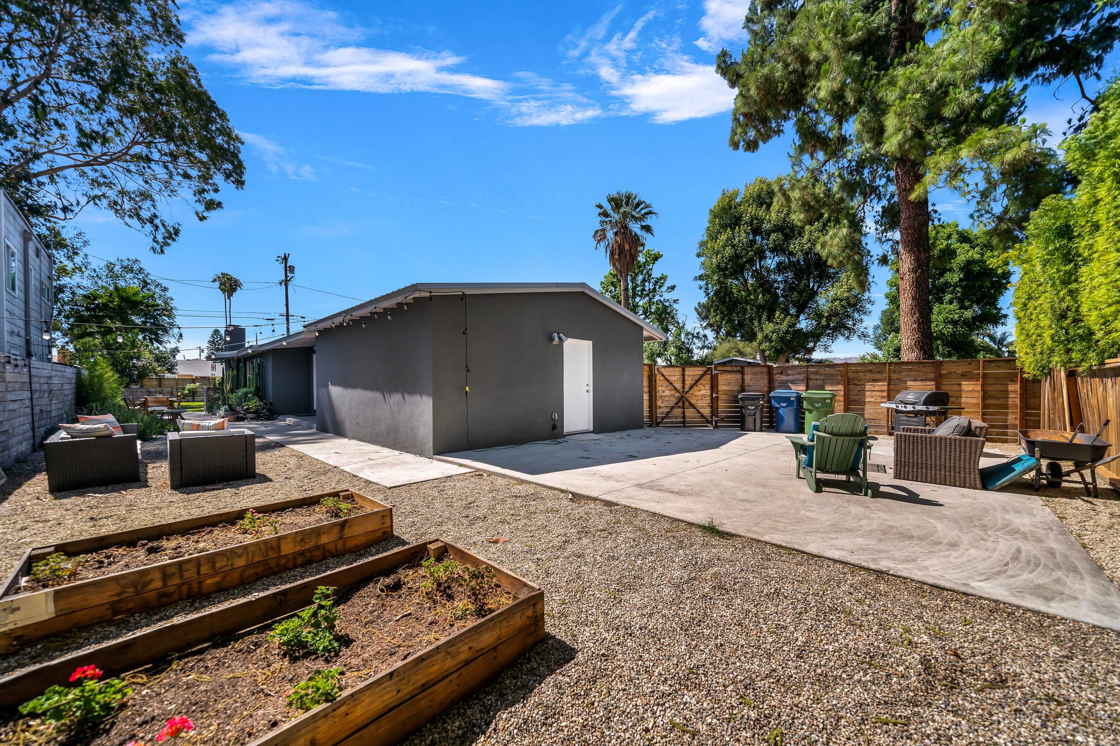 The image shows a spacious backyard with a gravel surface, raised garden beds, a gray building, and various outdoor furniture against a backdrop of trees.