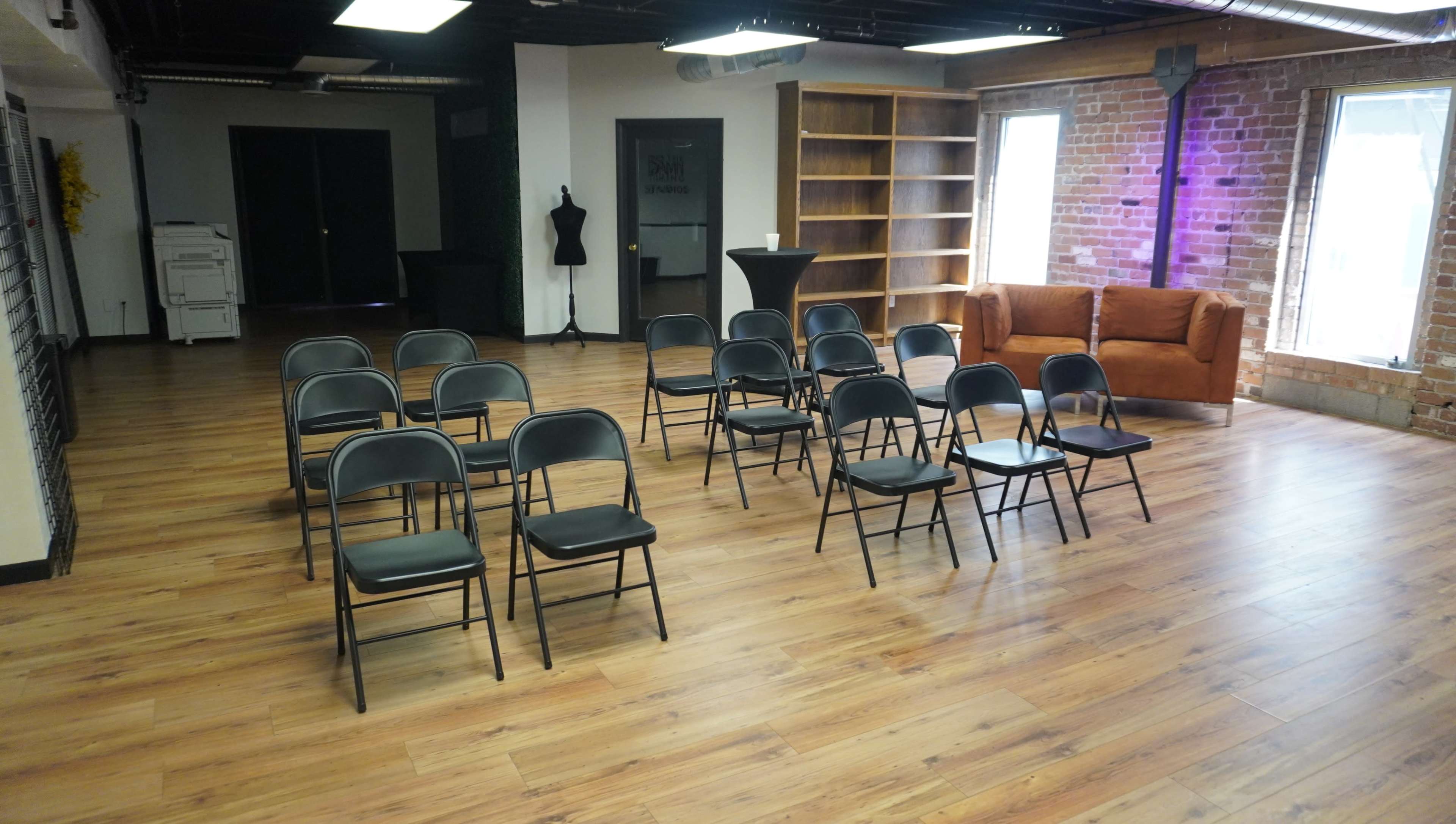 A row of black folding chairs is arranged in a partially empty room with wooden flooring and large windows, alongside a brown couch and a bookshelf.