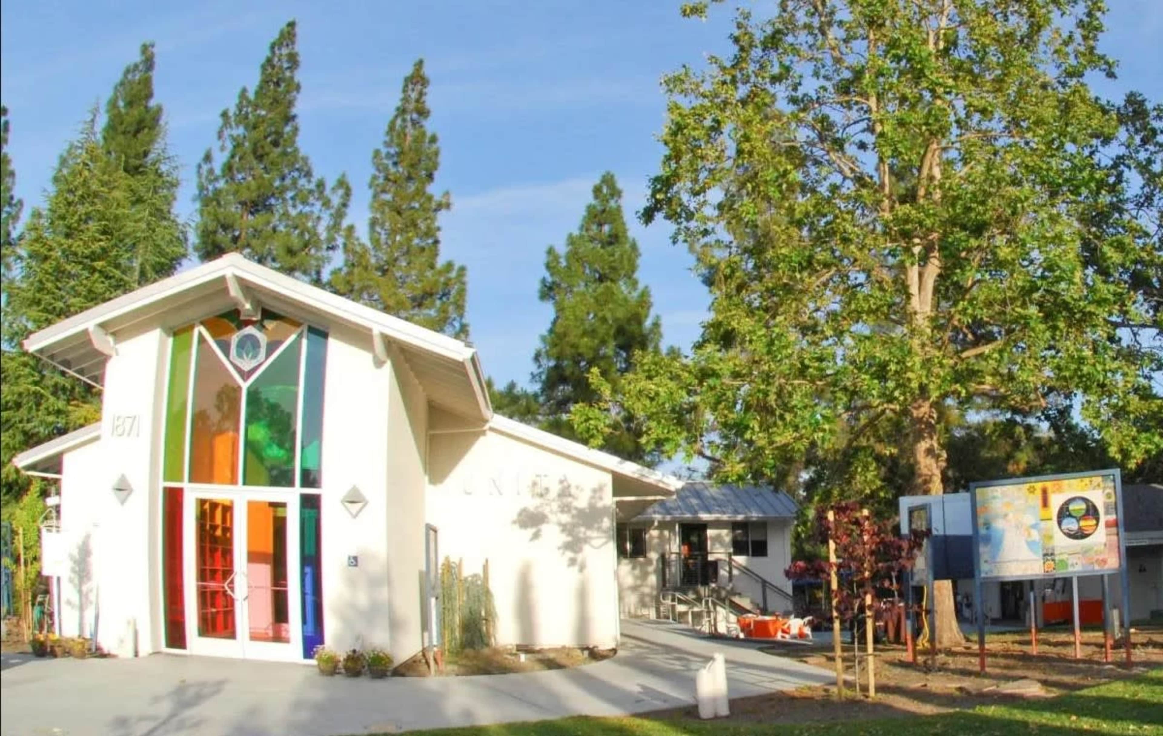 The image shows a modern building with large colorful windows, surrounded by green trees and a nearby information board.