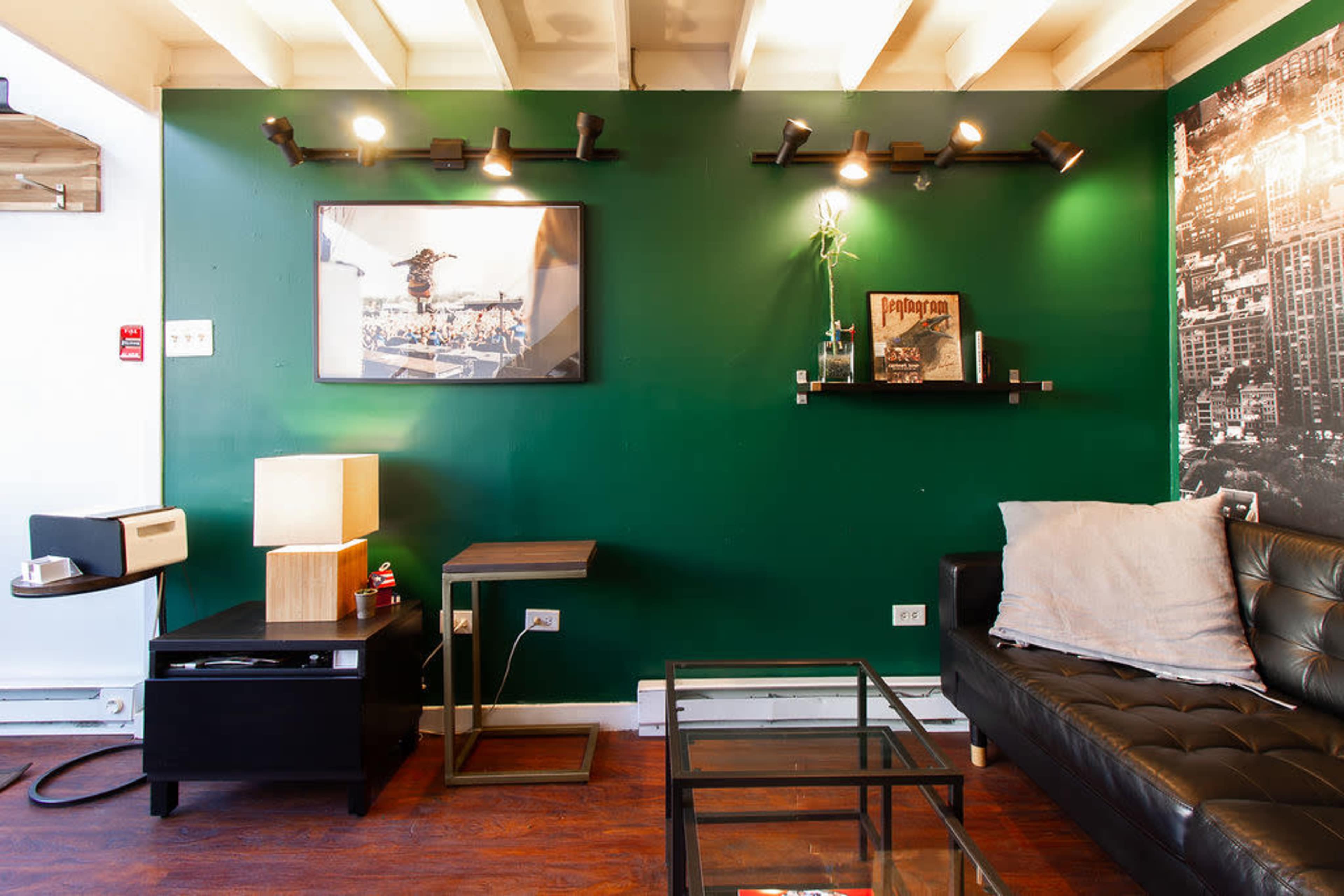 The image shows a modern living room with a green accent wall, featuring a black leather sofa, a glass coffee table, and a wall-mounted shelf with decorative items.