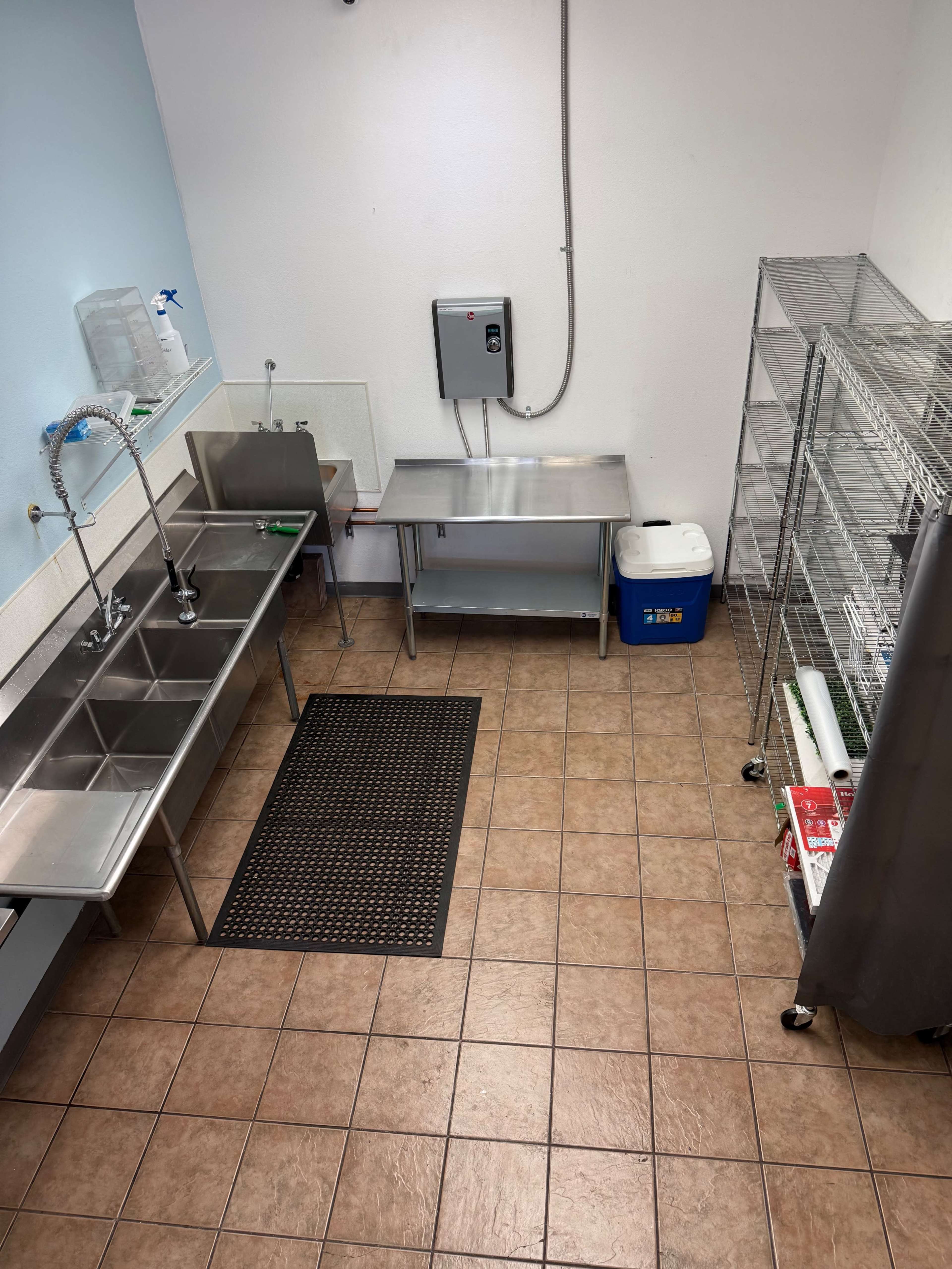 The image shows a commercial kitchen area featuring stainless steel sinks, a work table, a blue cooler, and metal shelving units against the wall.