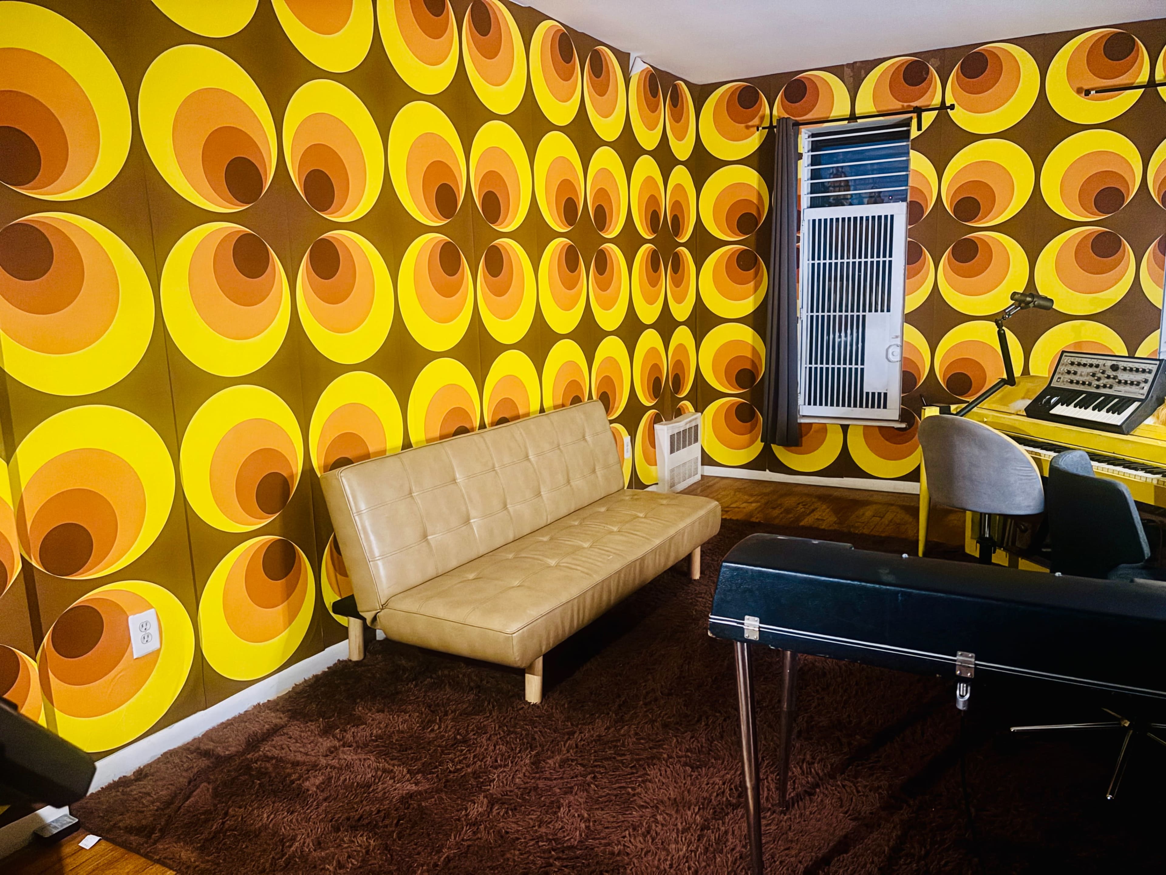 The room features a brown and orange patterned wallpaper, a tan sofa, a plush rug, and a keyboard setup in the corner.