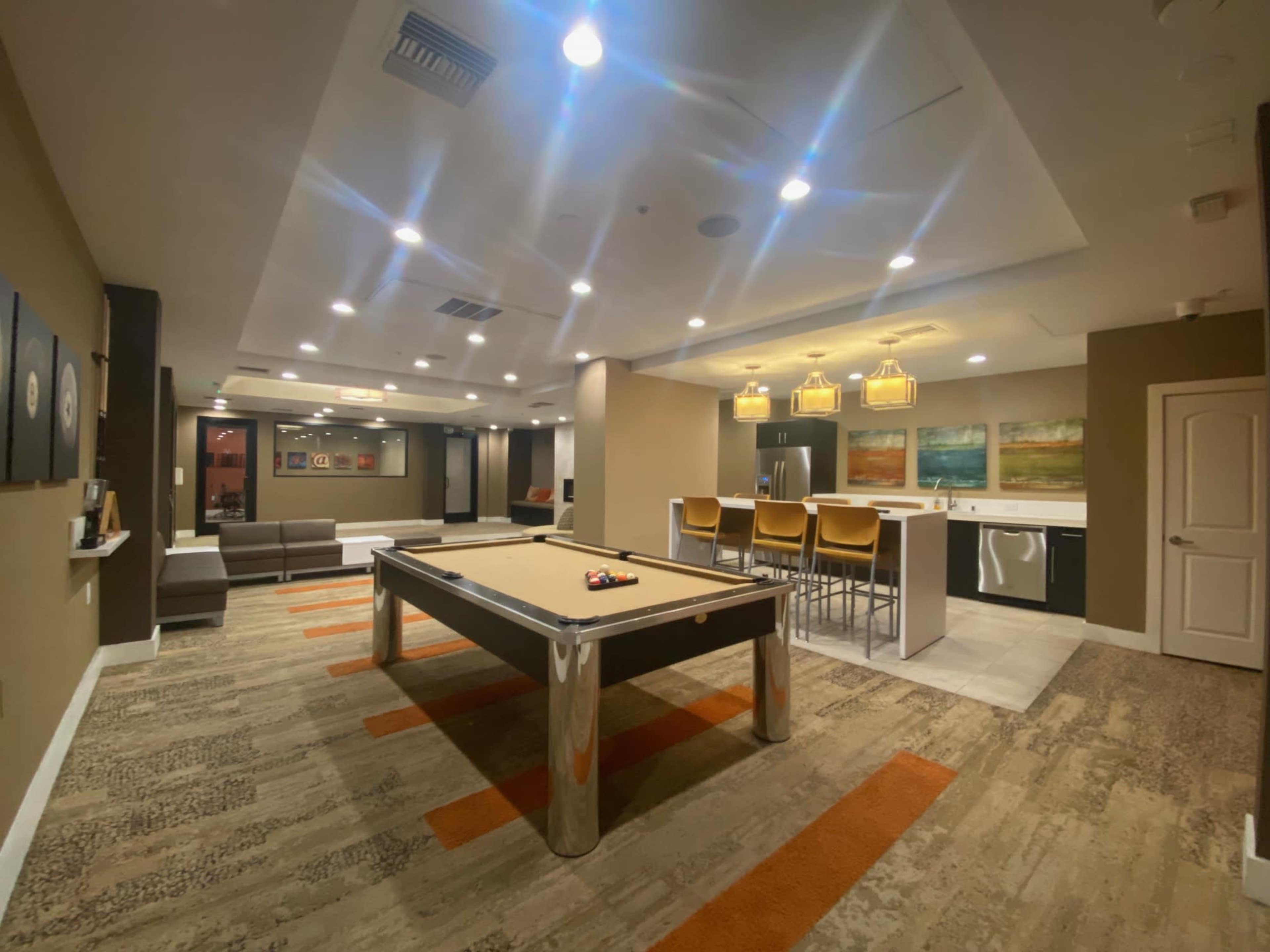 The image shows a recreational room featuring a pool table, seating areas, and a kitchenette with bar stools under pendant lights.
