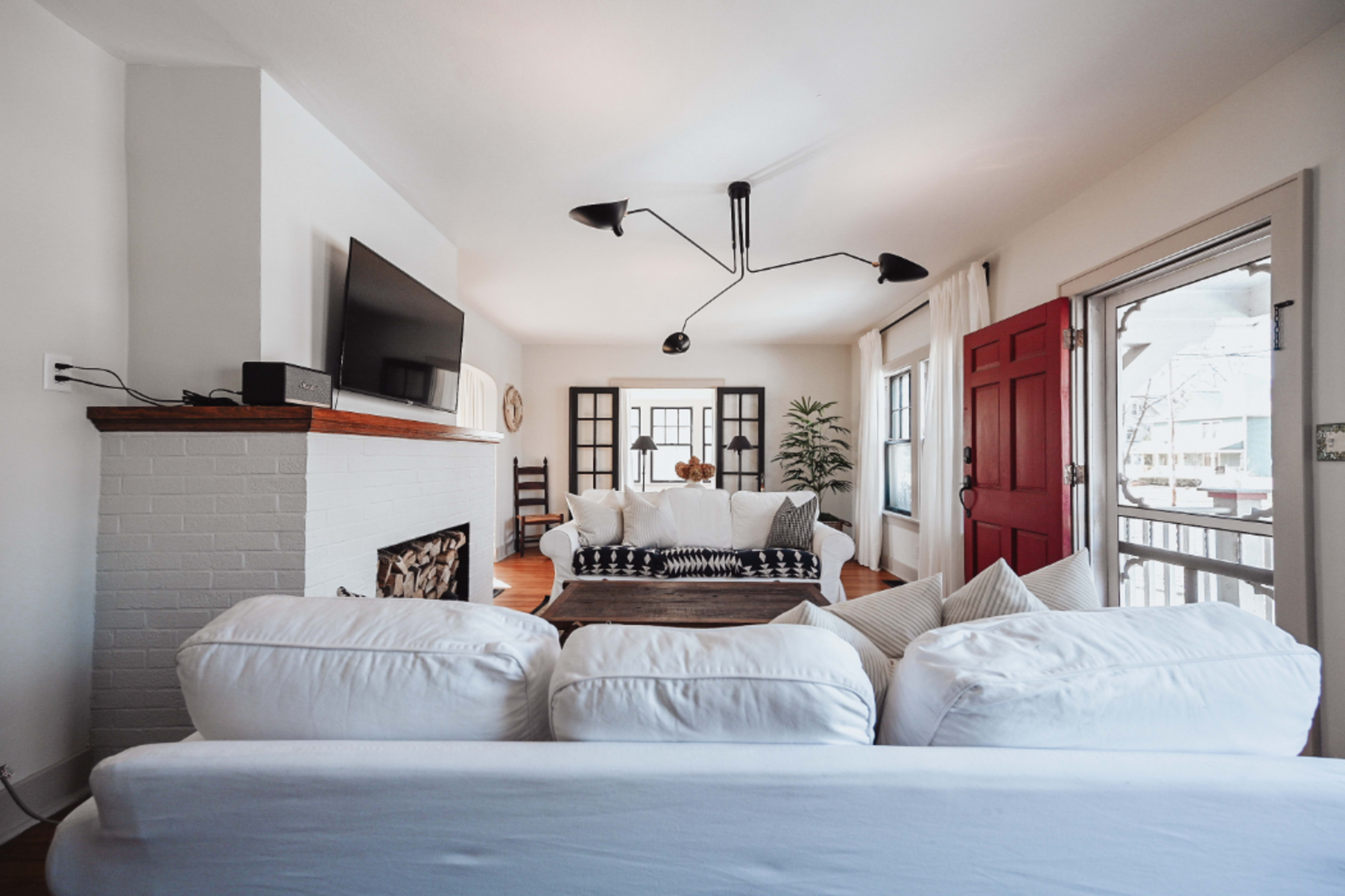 A spacious living room features a white sofa, a black-and-white patterned rug, a television mounted on the wall, and a bright red door leading outside.
