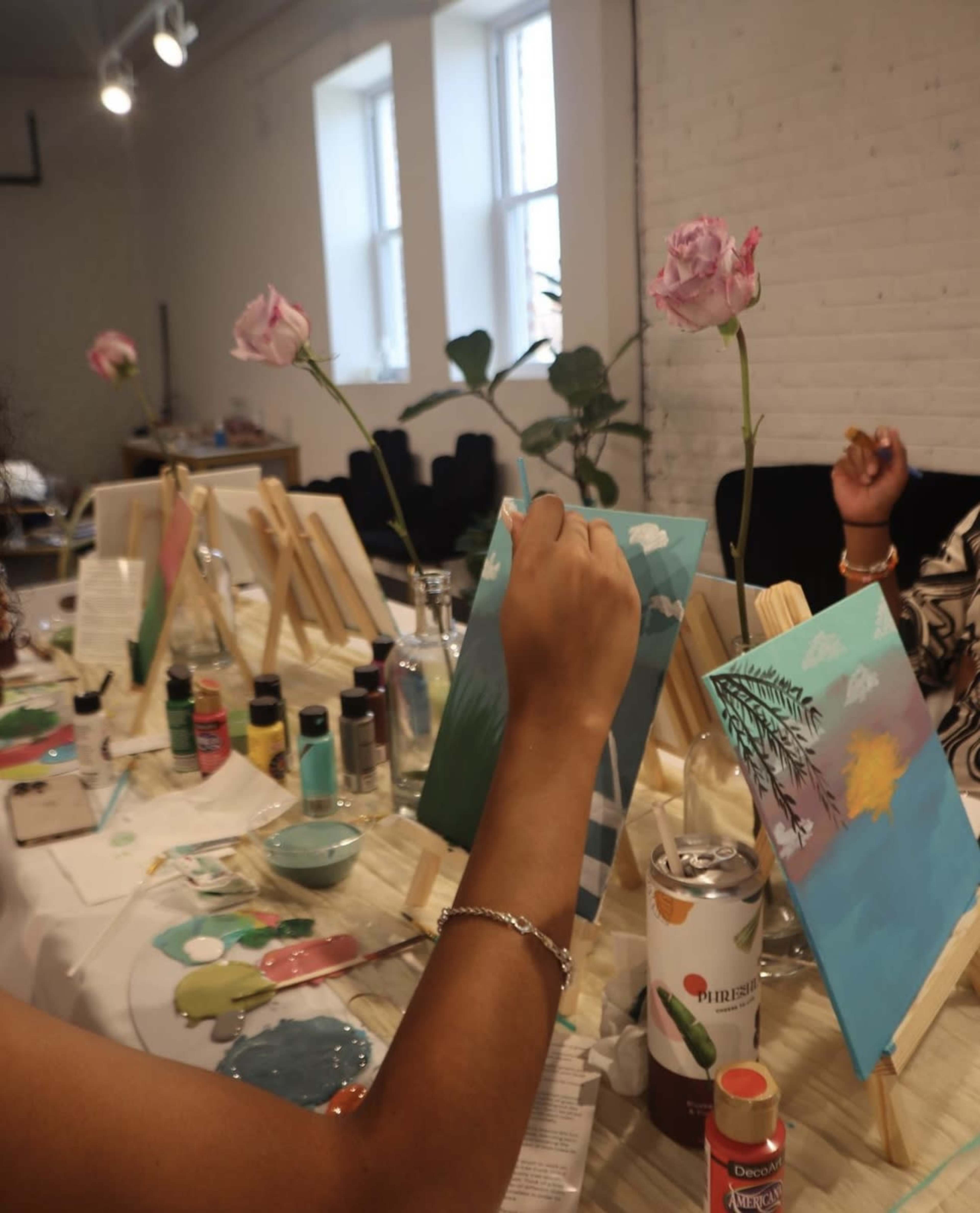 A person is painting on a small canvas while surrounded by other canvases, paint supplies, and fresh roses in vases.