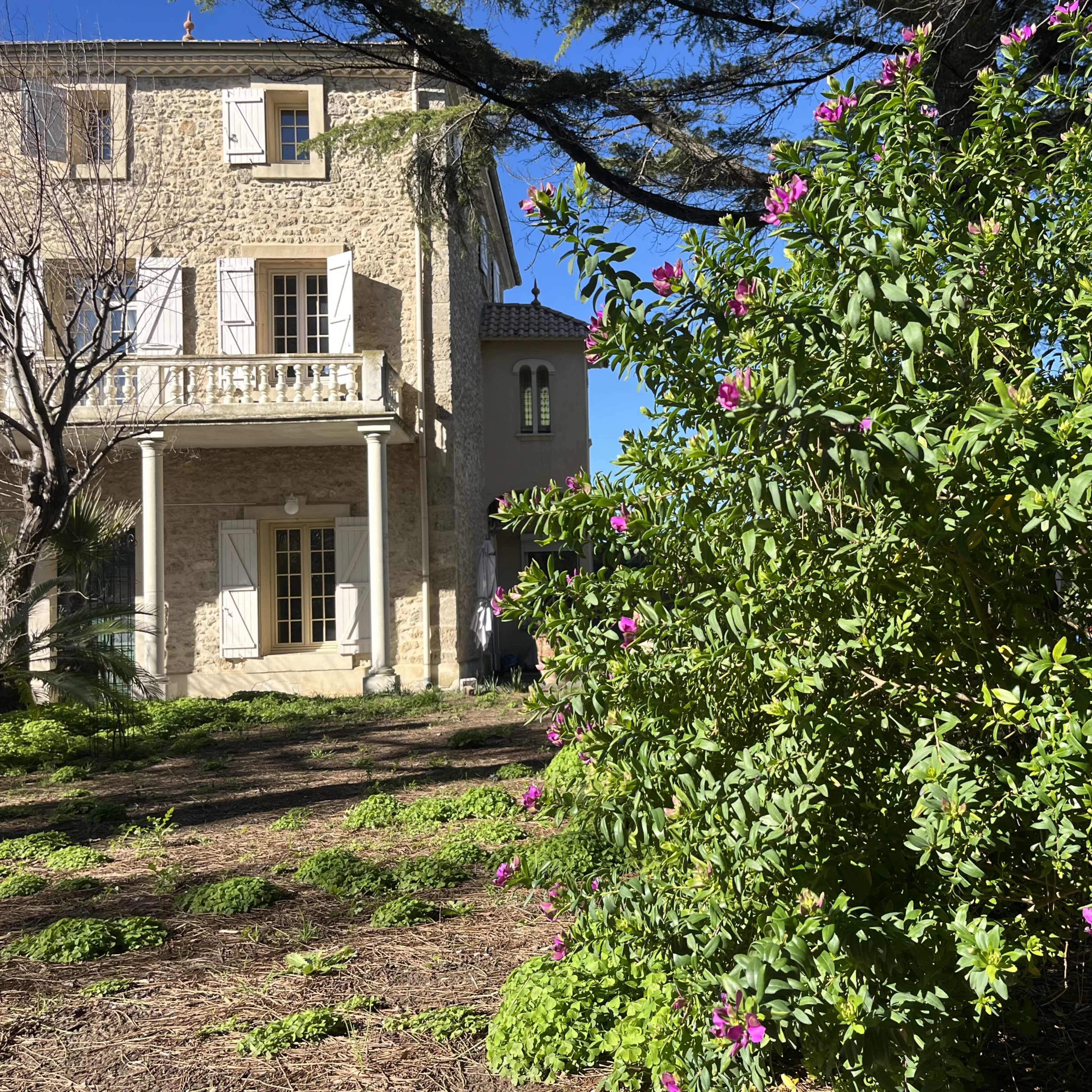 A stone house with white shutters stands amidst a garden featuring green foliage and pink flowers.