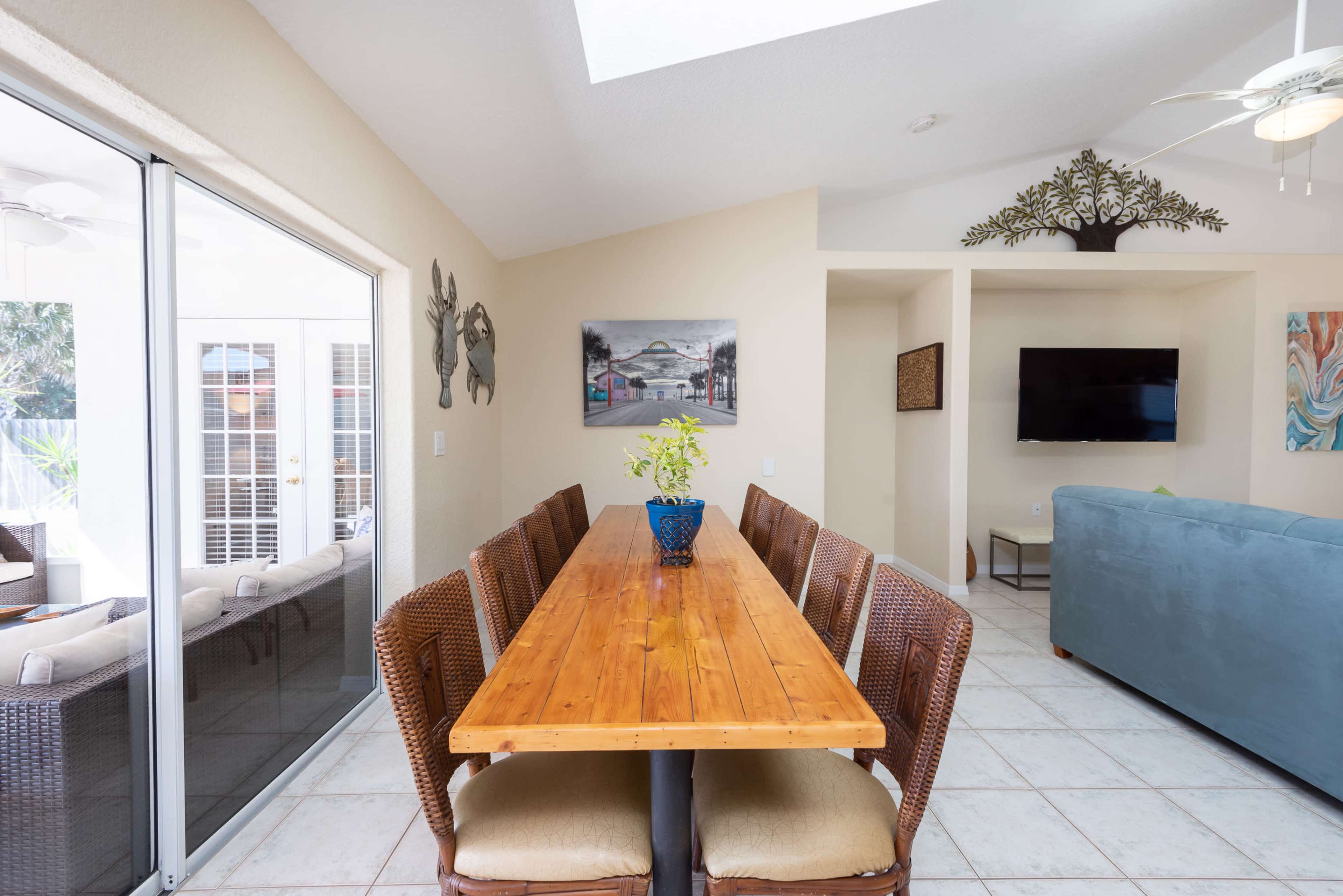 A long wooden dining table with six chairs is set in a brightly lit room featuring a large window and a wall-mounted television.
