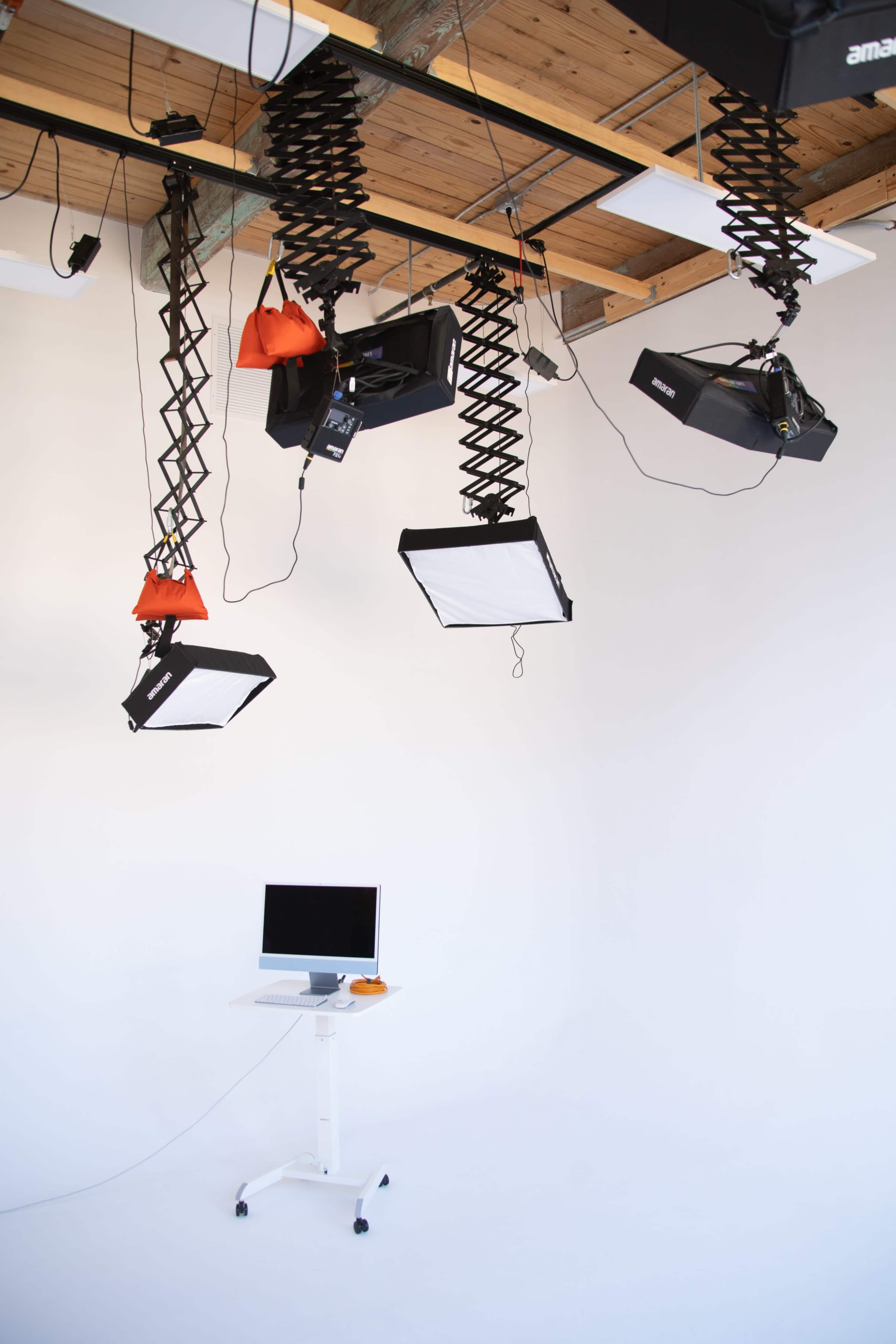 The image shows a photography studio with multiple overhead lights and a computer on a mobile stand against a white backdrop.