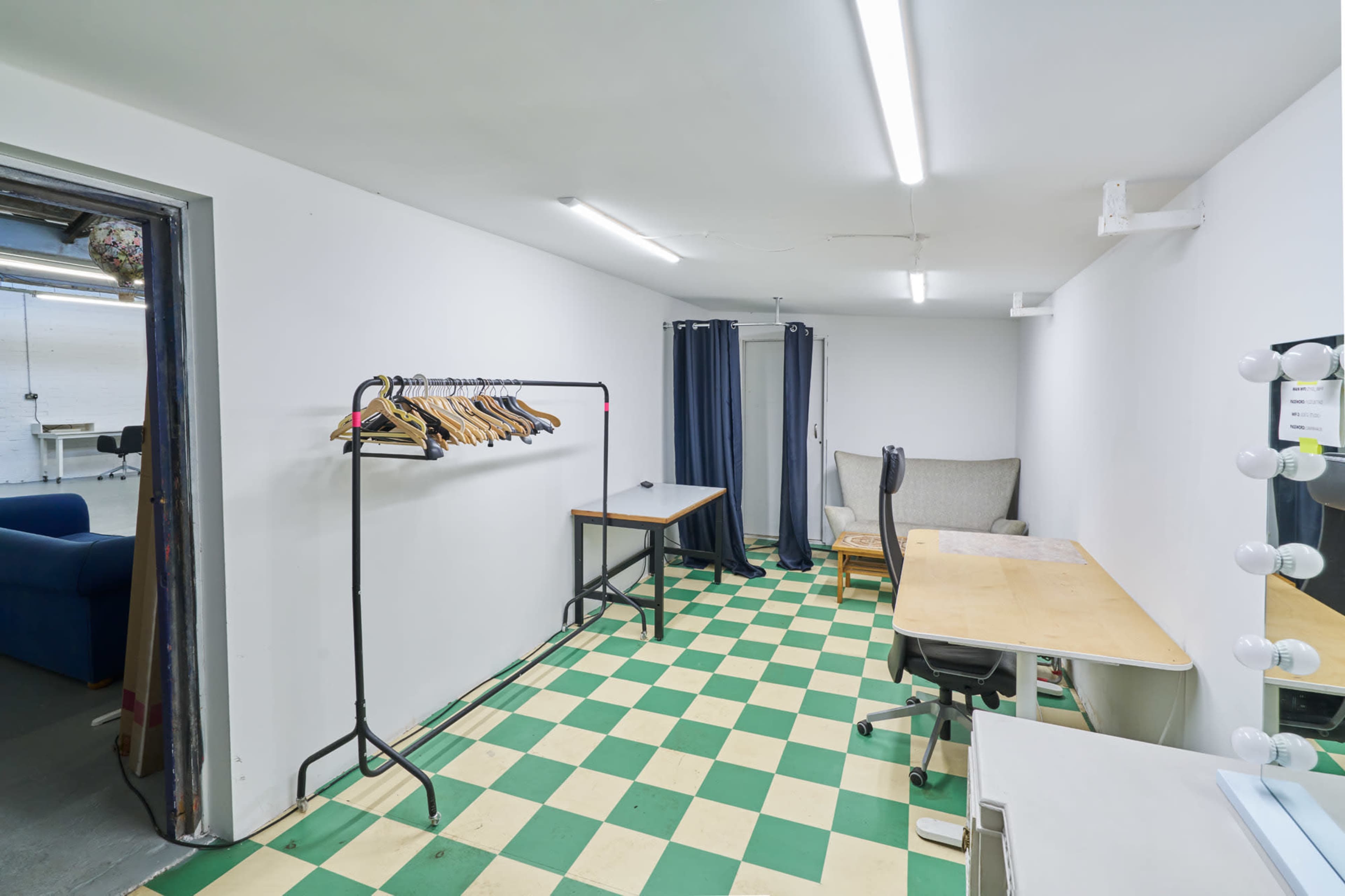 A small, sparsely furnished room features a clothing rack, a table with chairs, and a large mirror surrounded by lights, with checkered flooring and plain white walls.
