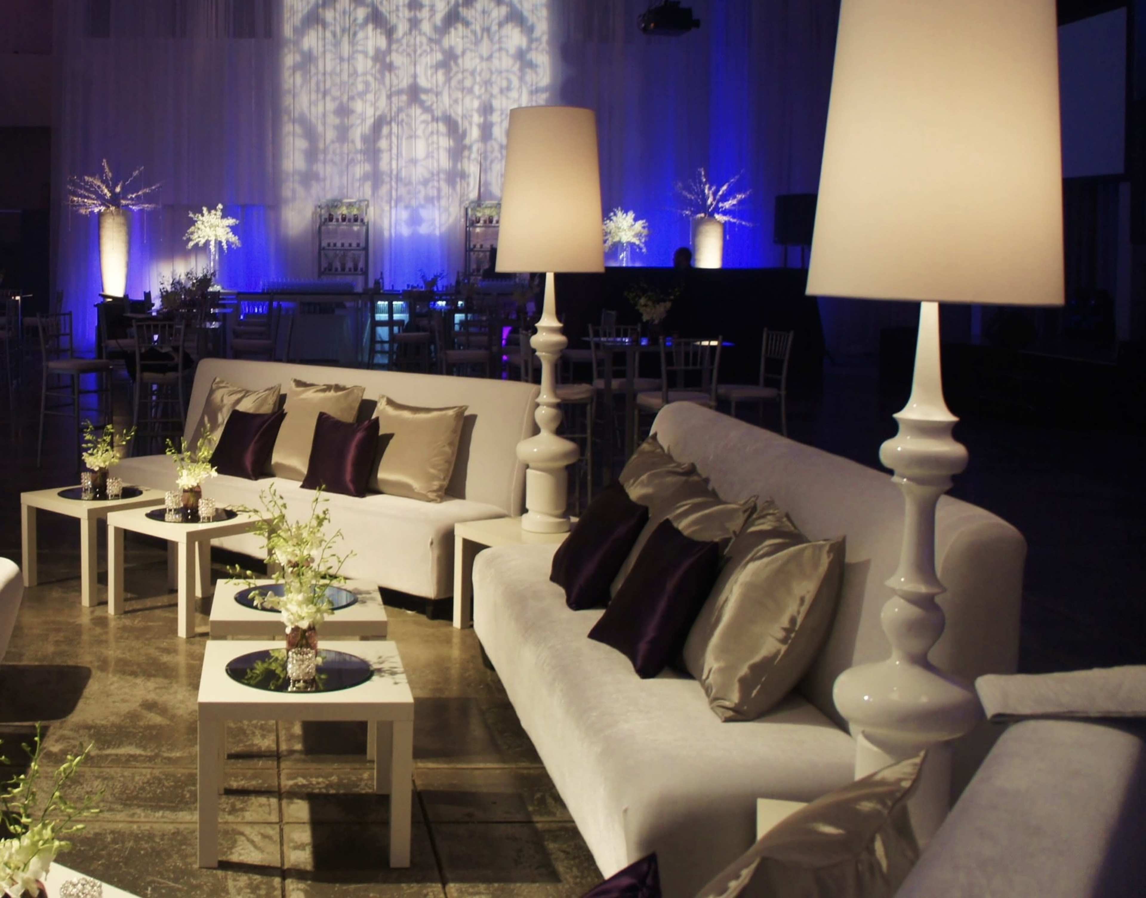 The image shows a stylish event space featuring white sofas with purple and gold accents, modern lamps, and small tables with floral arrangements.