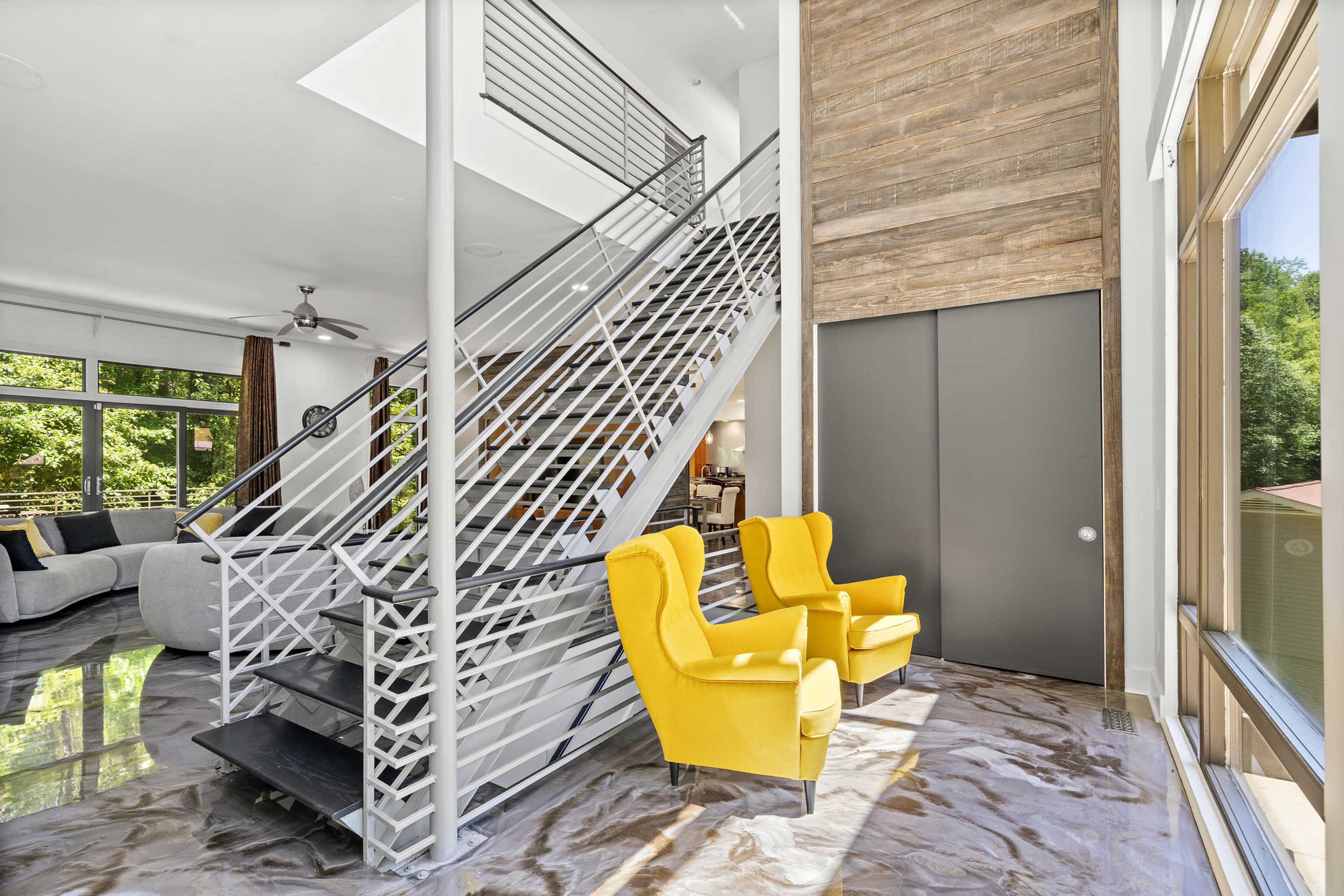The image shows a modern interior featuring a staircase with a metal railing, two yellow armchairs, and large windows that provide natural light.