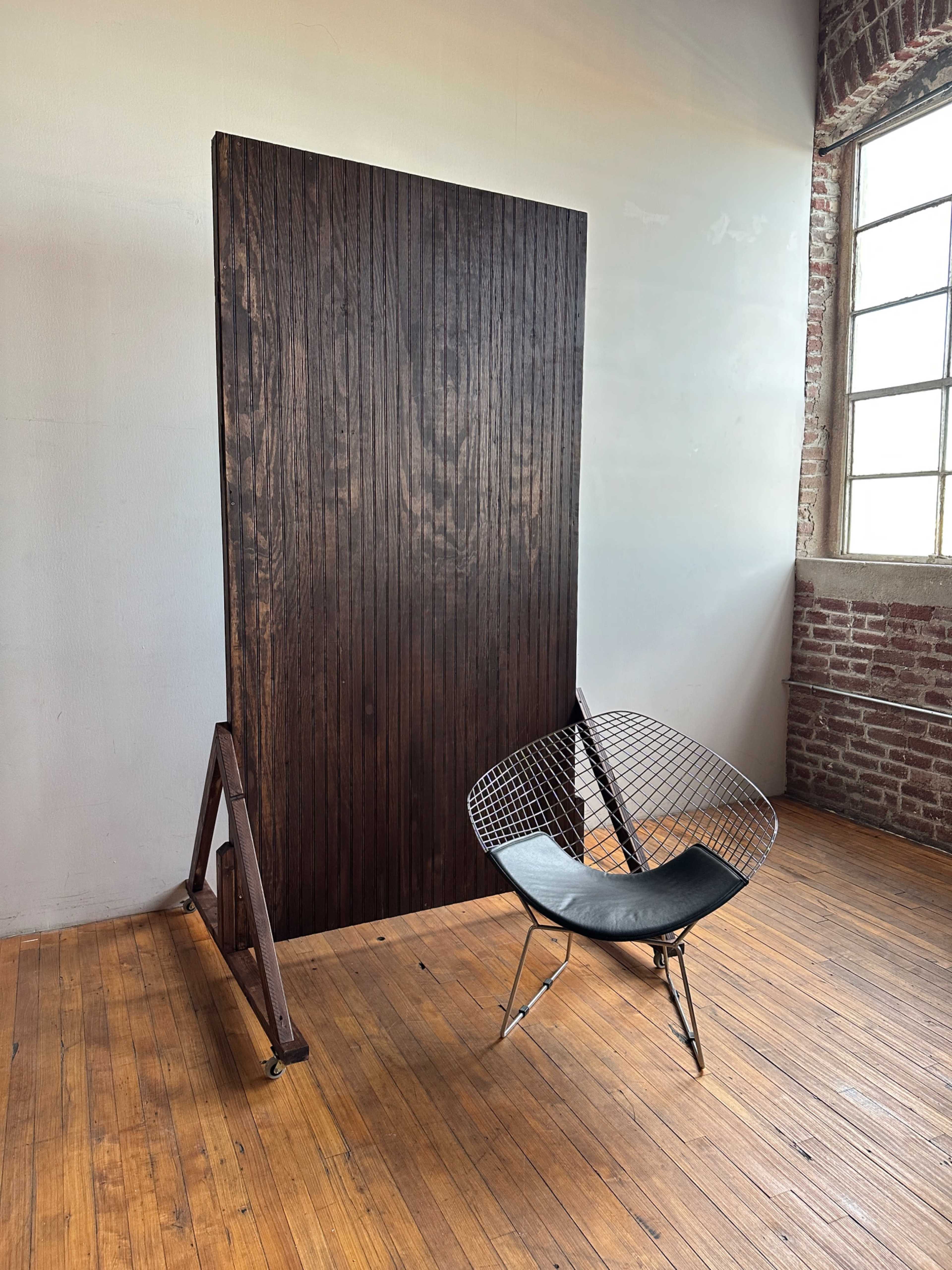 A large wooden panel stands against a wall next to a modern wire chair on a wooden floor.