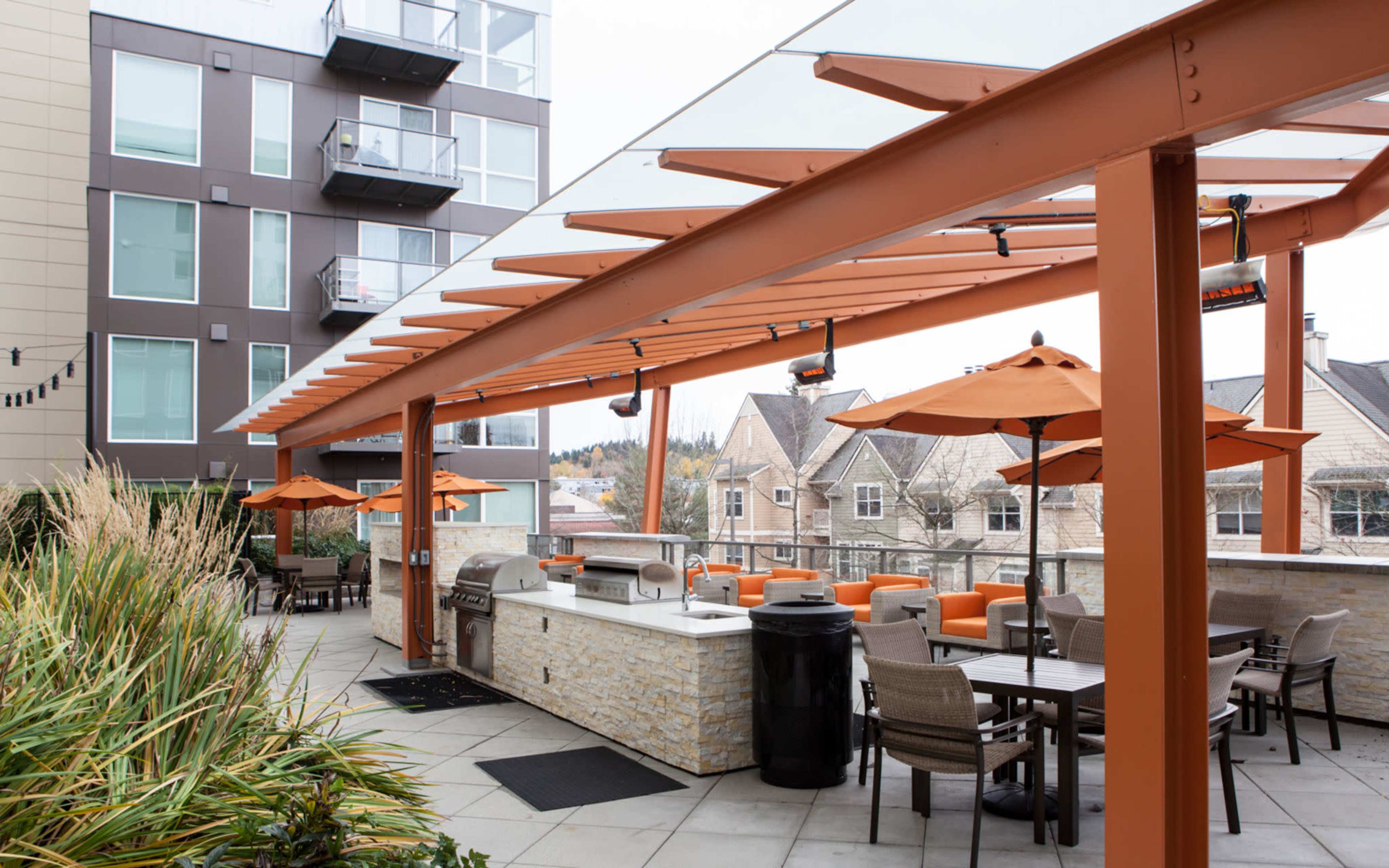 An outdoor patio area features orange umbrellas, seating arrangements, and a grill under a canopy with an apartment building in the background.
