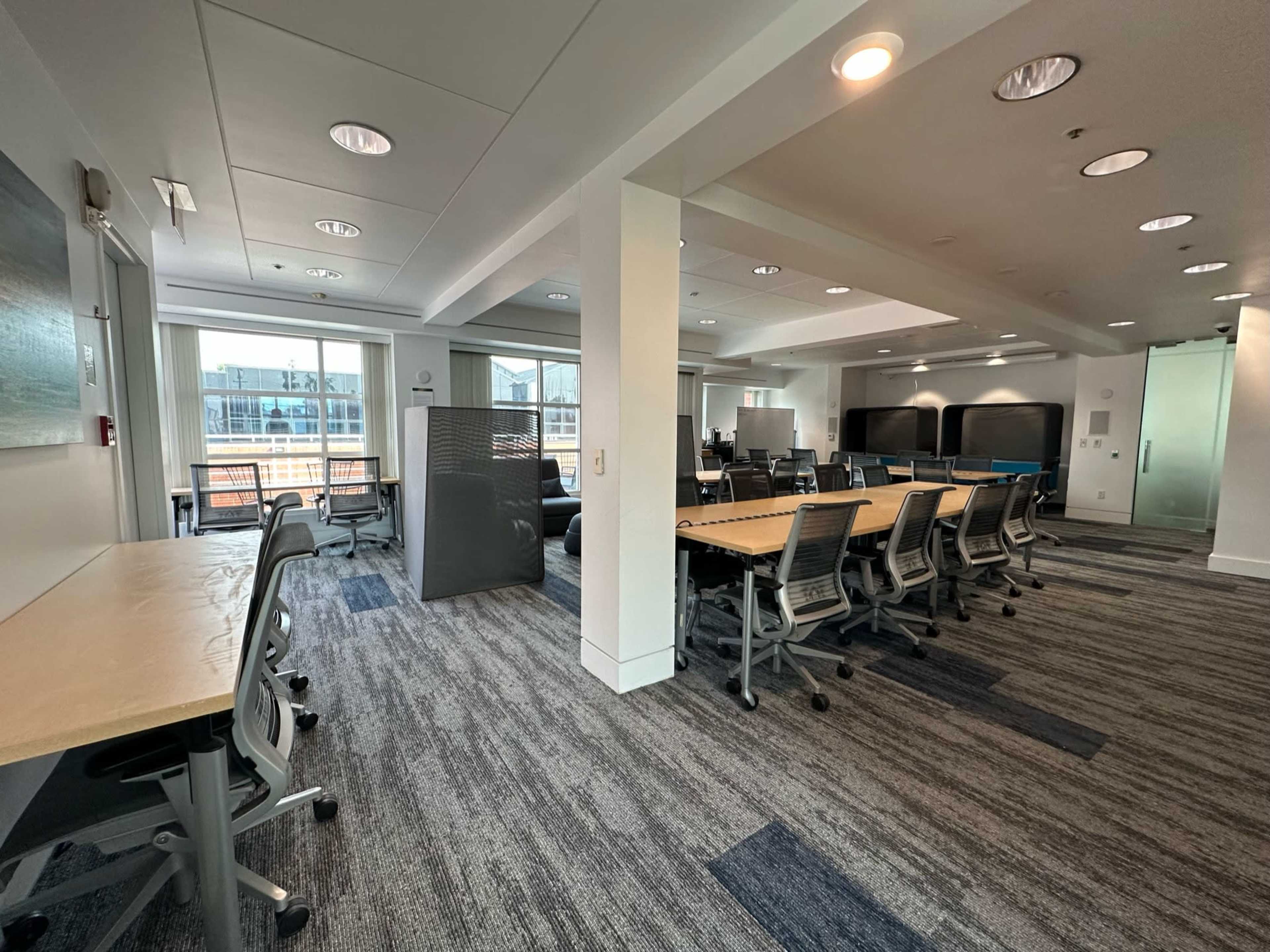 An office space featuring several desks and chairs arranged in a brightly lit room with large windows and modern furnishings.