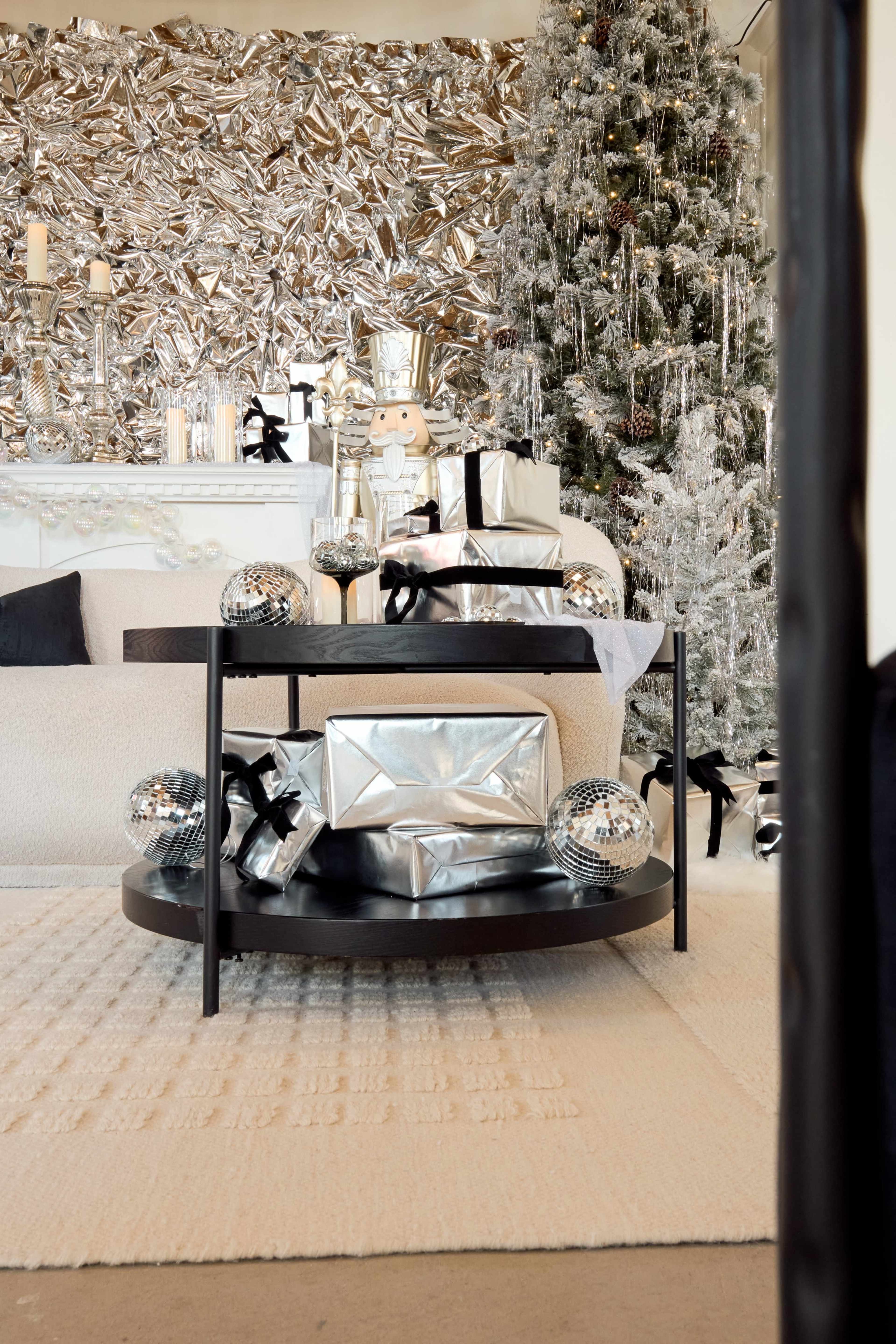 The scene features a modern living room decorated for the holidays, with a sleek coffee table displaying silver-wrapped gifts and disco balls, set against a backdrop of shiny silver wall decor and a Christmas tree.