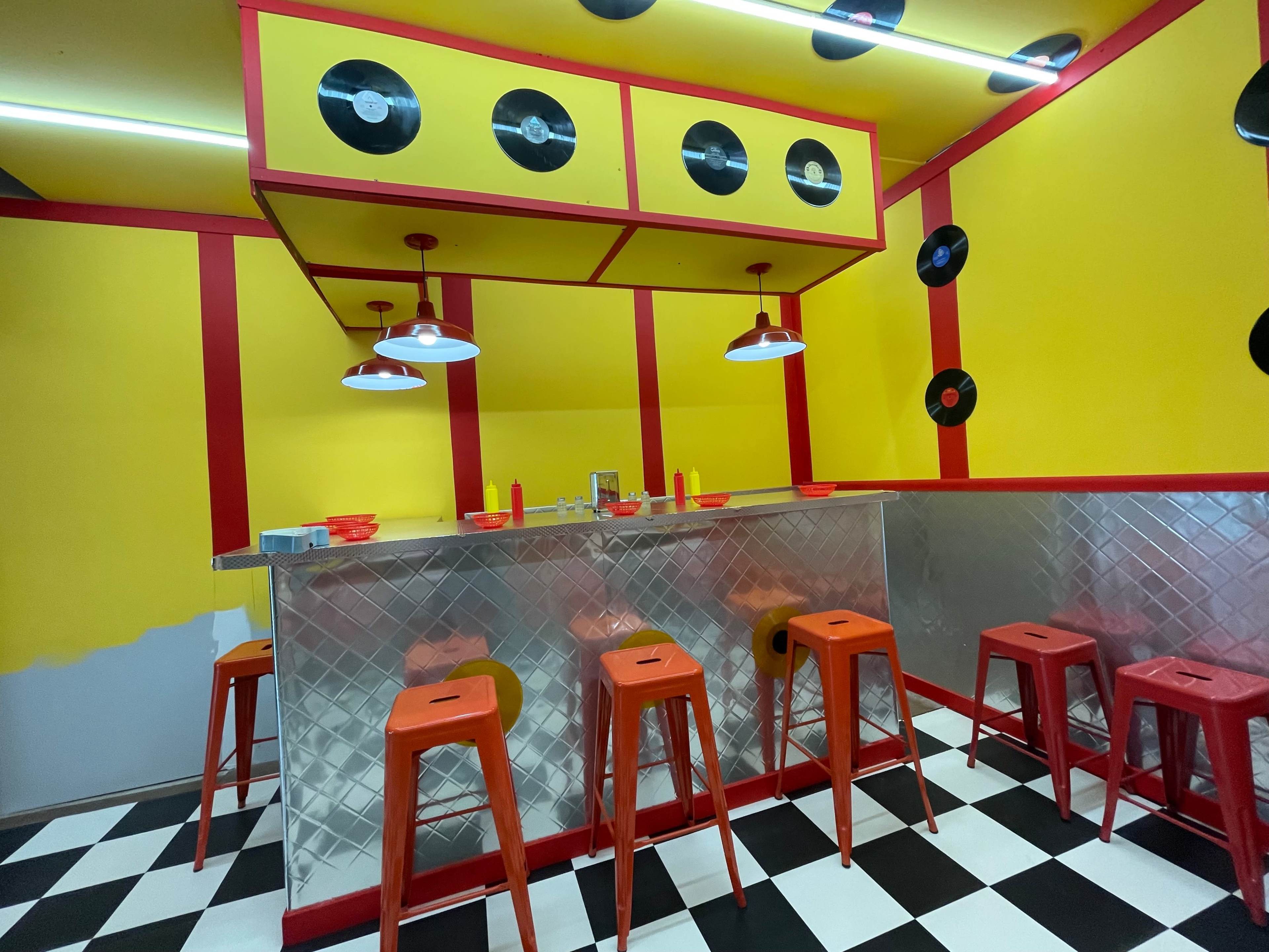 A retro-themed diner features a checkered floor, yellow walls adorned with vinyl records, and a silver counter with red stools.