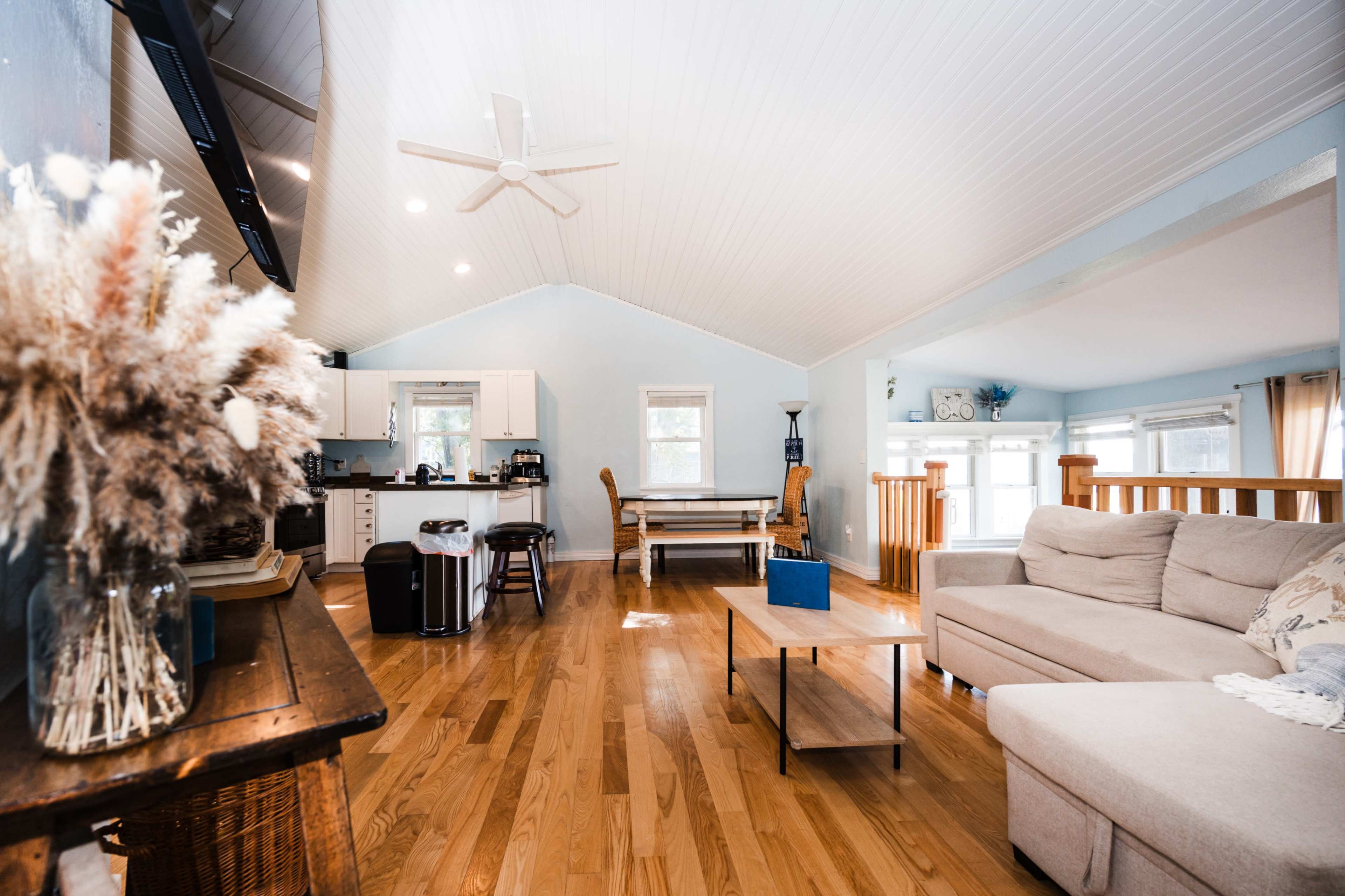 The image shows a spacious, well-lit living area with a light-colored sofa, wooden flooring, and a kitchen area visible in the background.