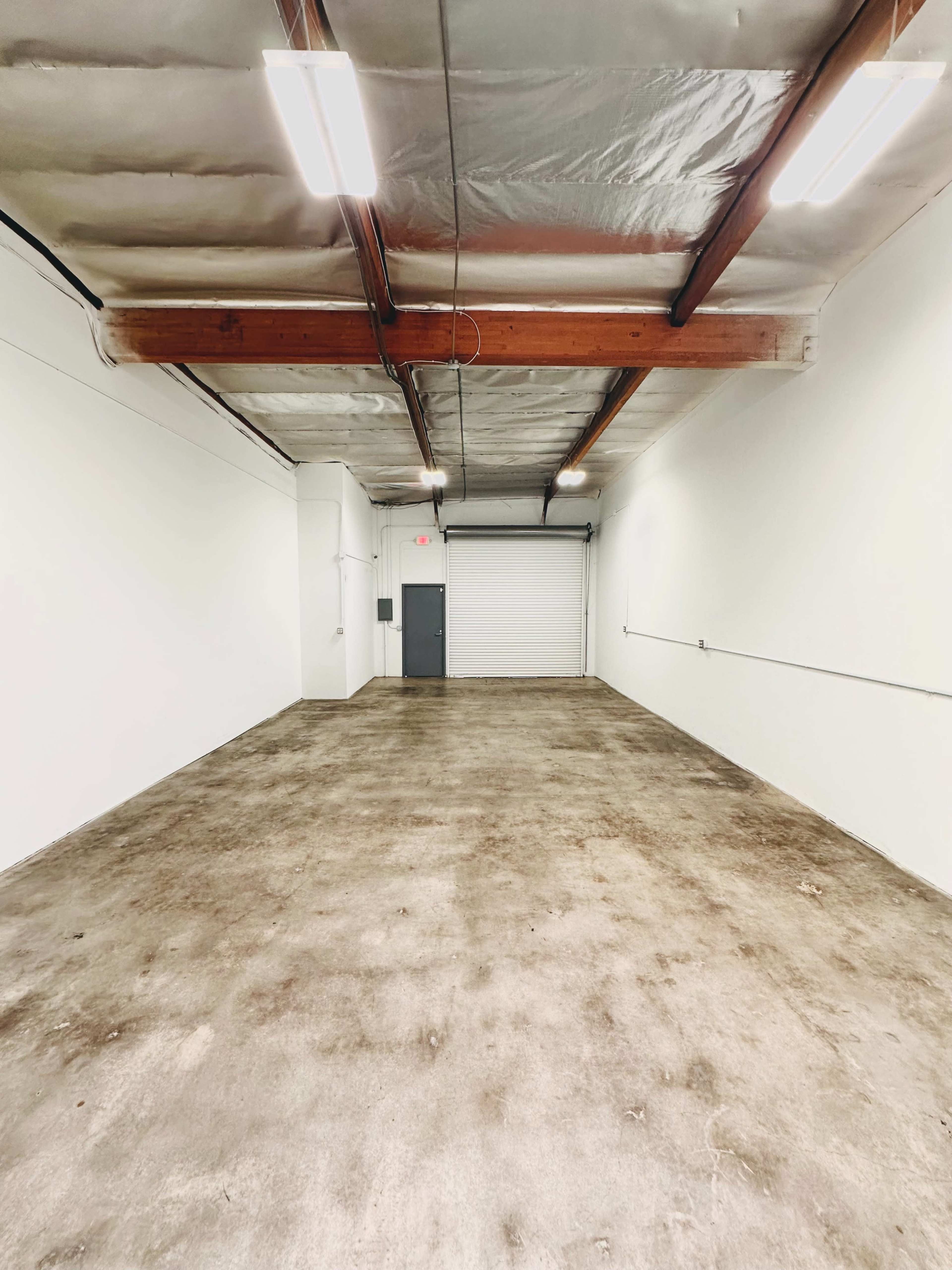 A large, empty garage space with a concrete floor and white walls, featuring a roll-up door at one end and exposed beams on the ceiling.