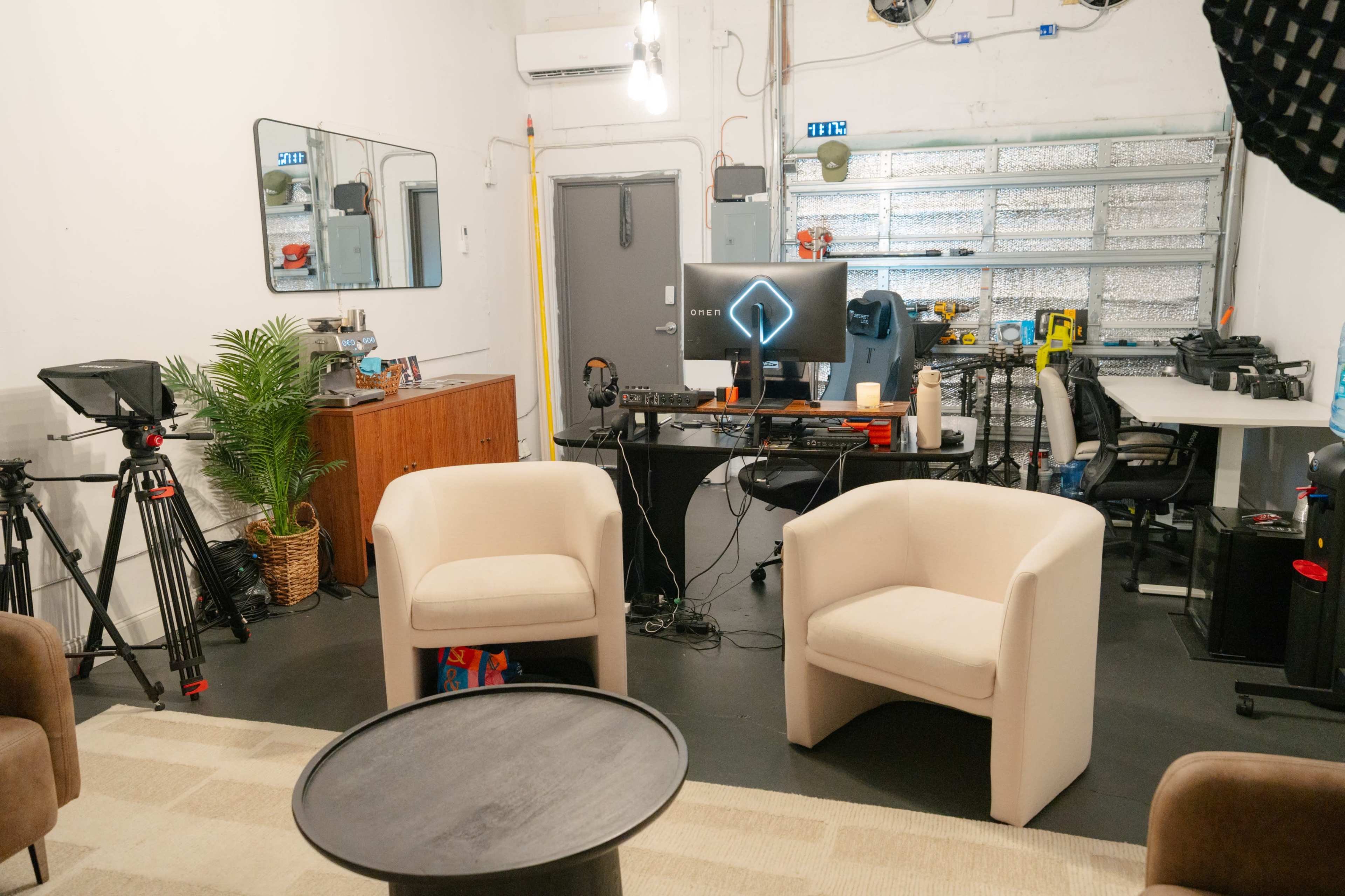 The image shows a small studio space with two white chairs facing a dark table, surrounded by various equipment including cameras and a computer station.