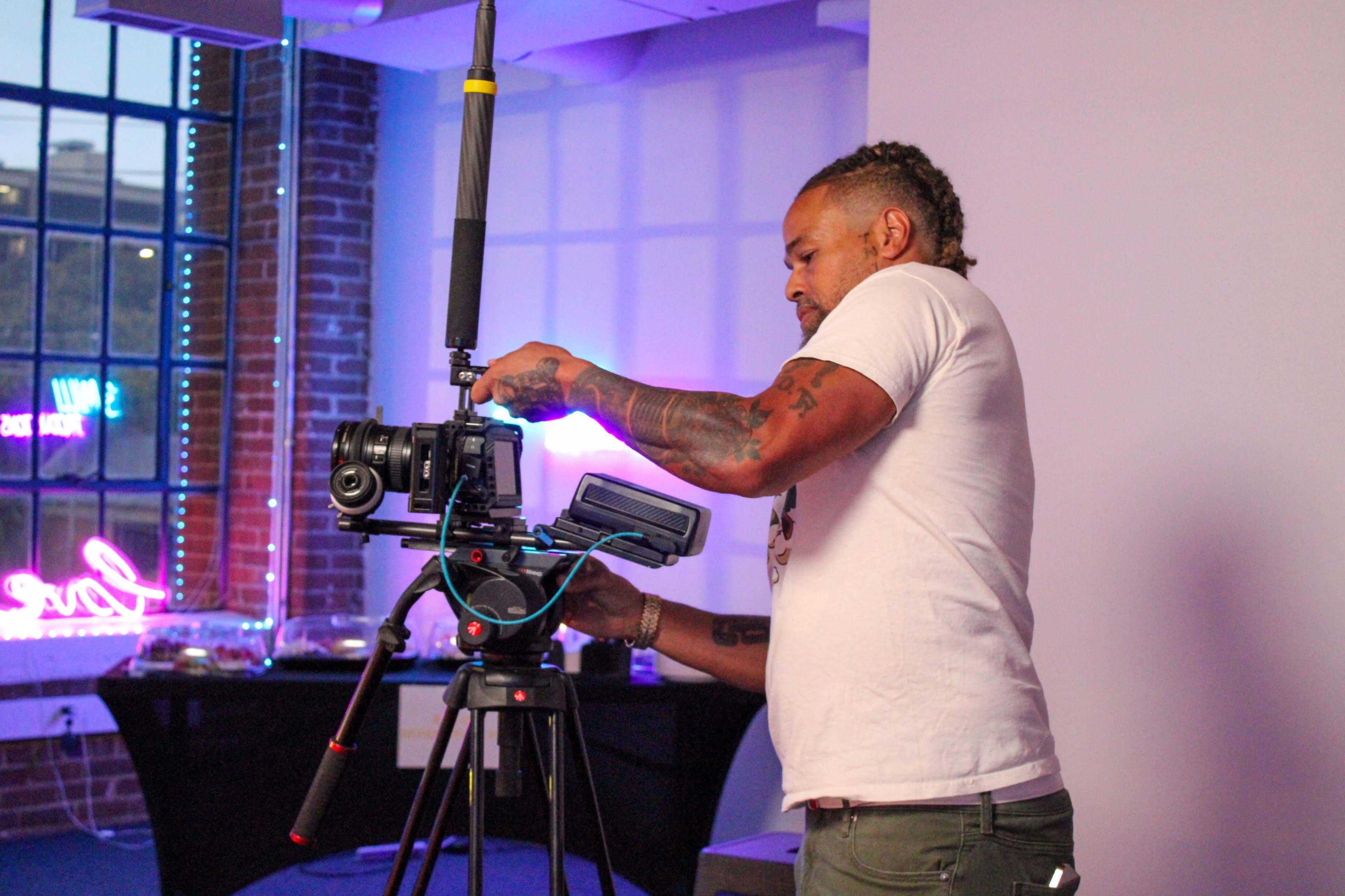 A man adjusts a camera setup on a tripod in a room with a large window and decorative lighting.