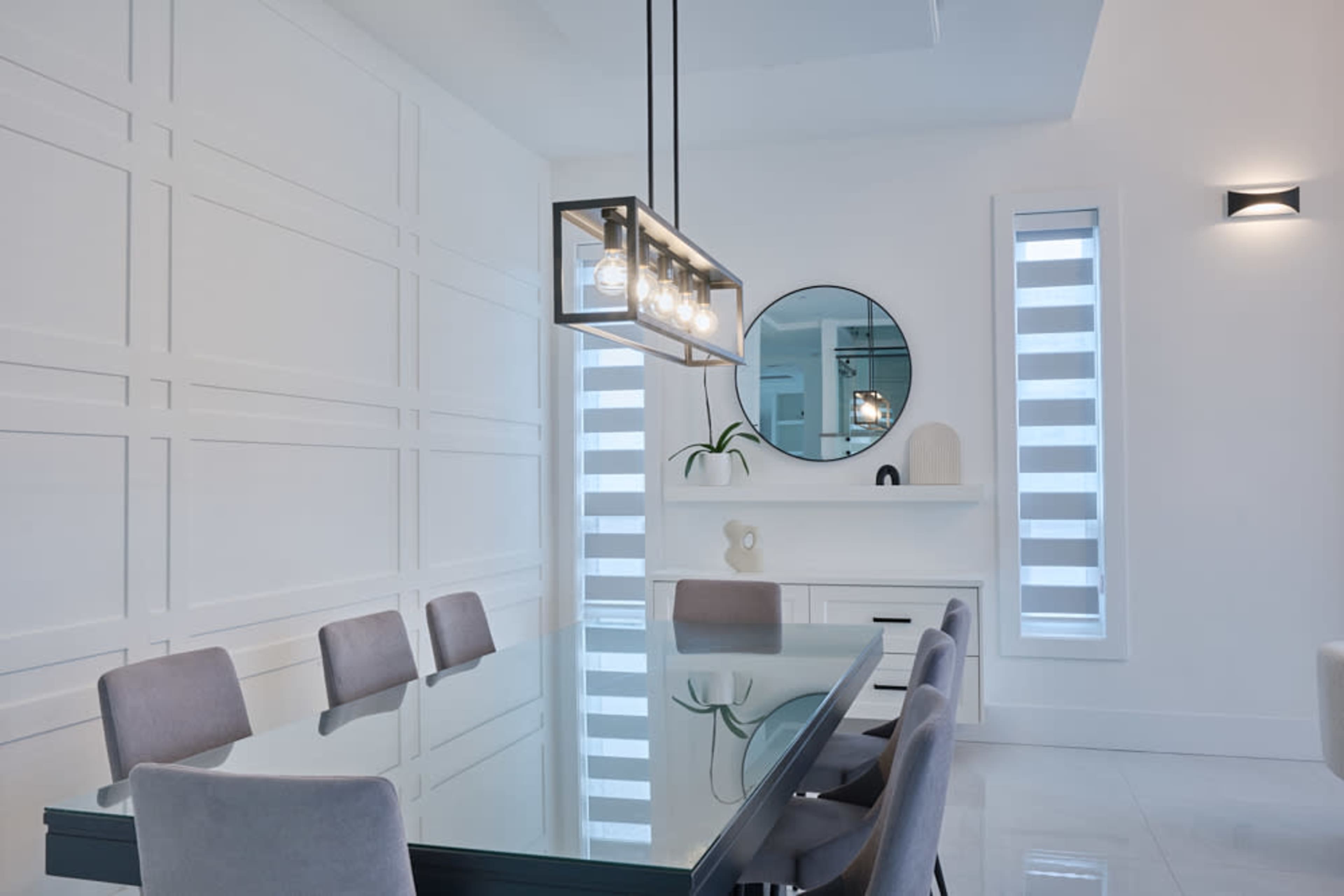 A modern dining room featuring a glass dining table with gray upholstered chairs, a round mirror, and a minimalist lighting fixture.