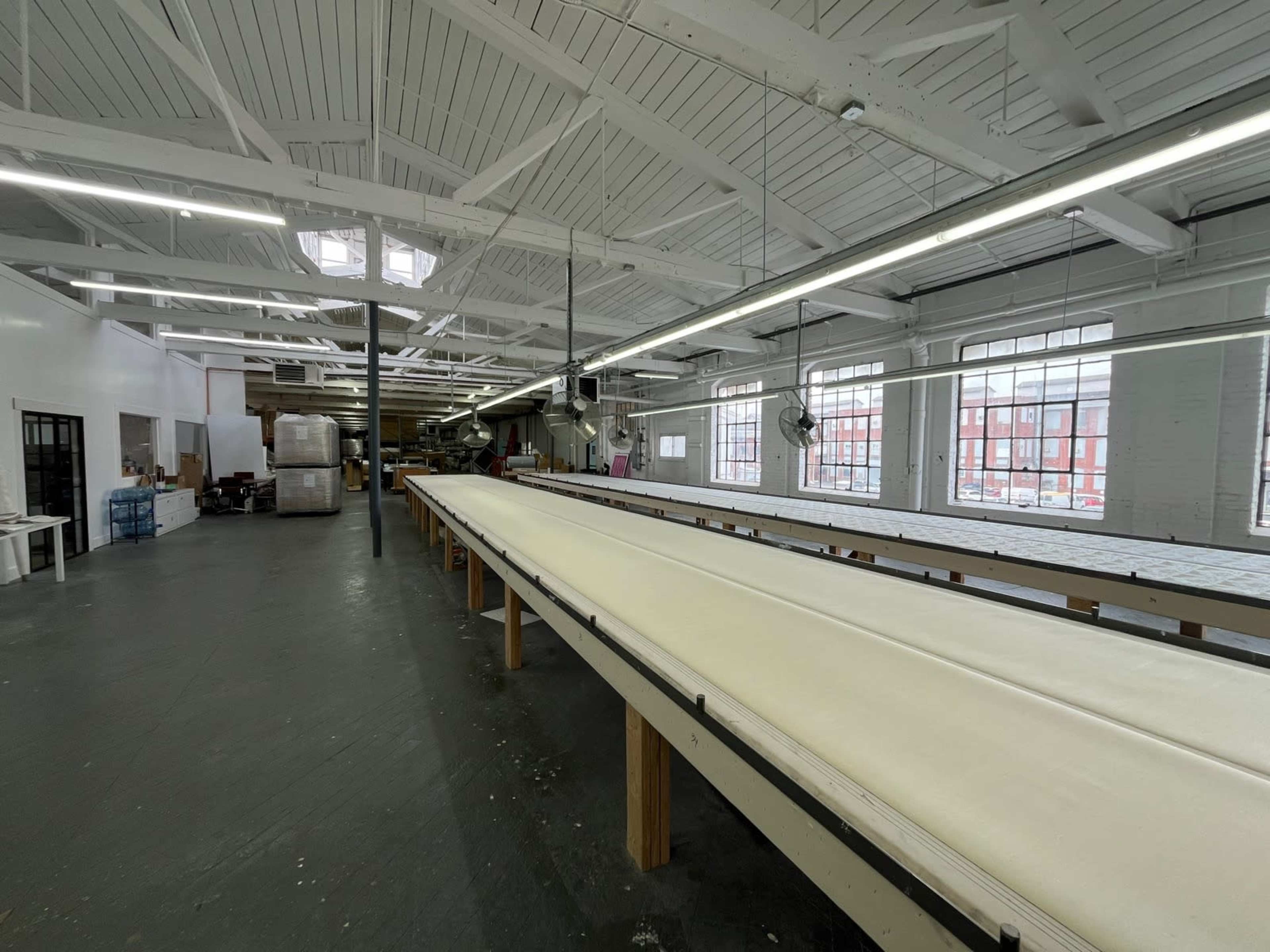 The image shows a spacious industrial workshop with long tables and large windows, illuminated by overhead lights.
