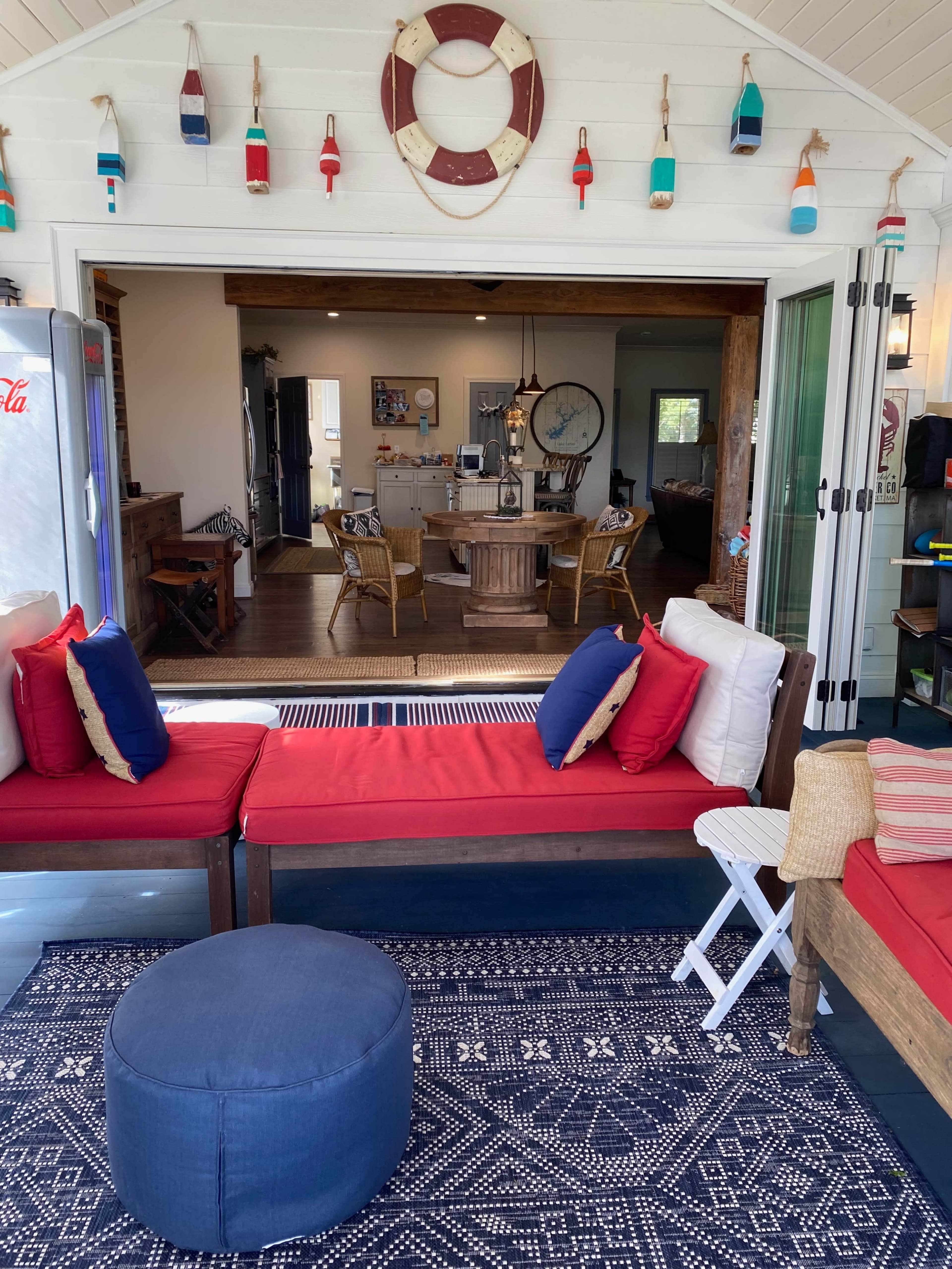 A cozy outdoor seating area with red cushions, a blue pouf, and a view of a well-decorated indoor space through open glass doors.