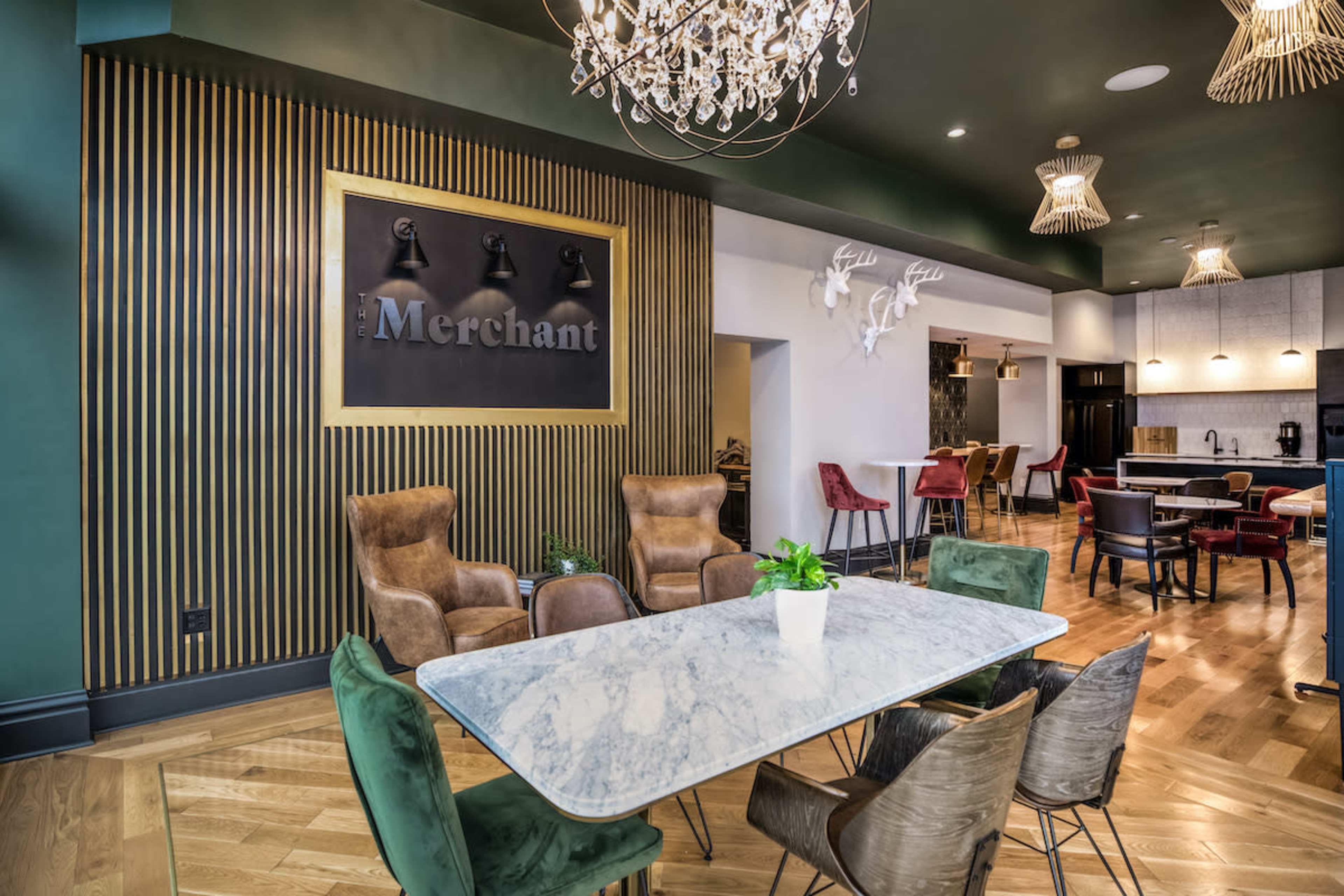 A stylish dining area featuring a large marble table surrounded by upholstered chairs, with a decorative wall displaying the word "Merchant" and accent lighting from chandeliers and wall-mounted fixtures.