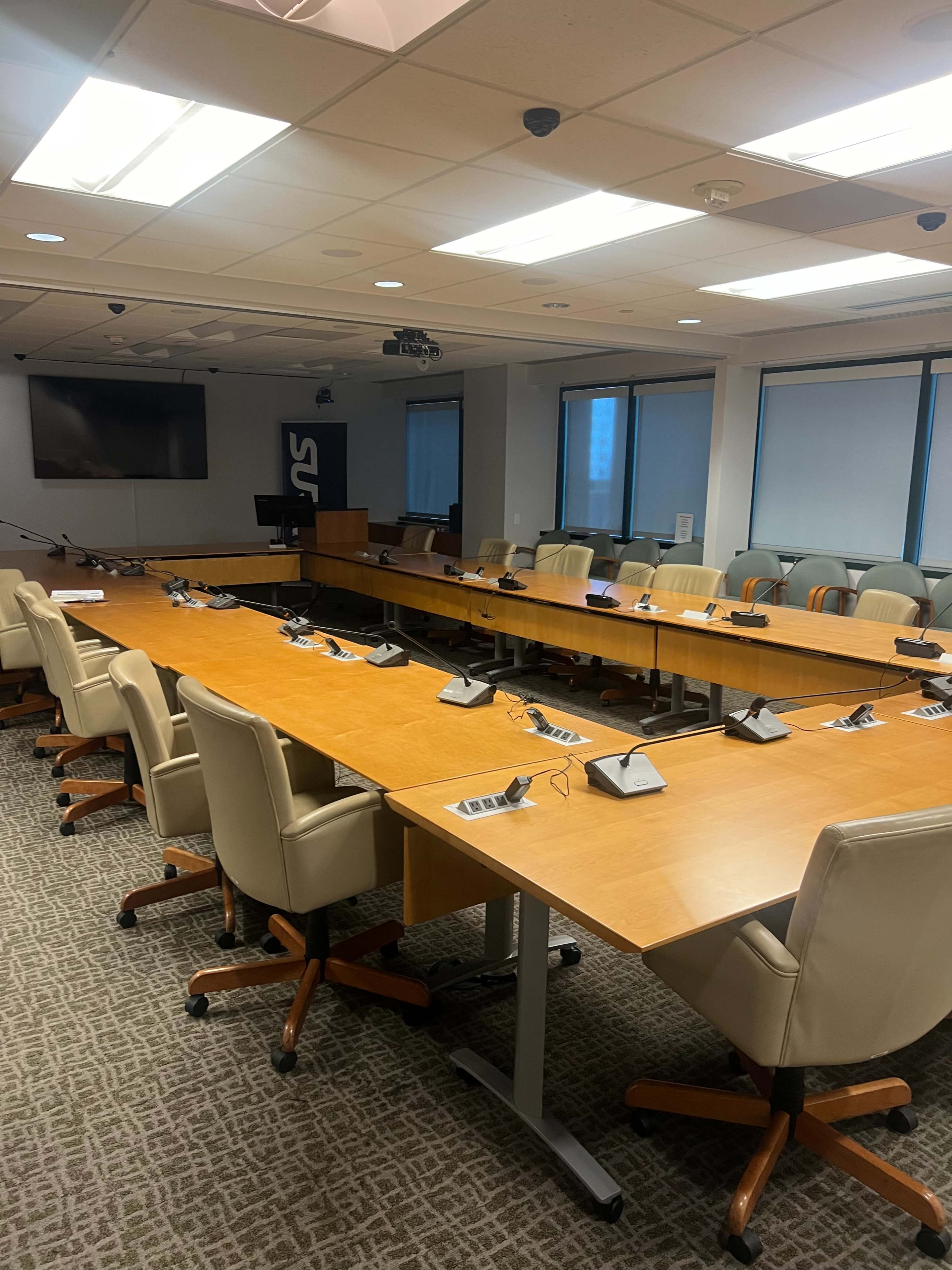 A large conference room features a long wooden table surrounded by office chairs and equipped with audio-visual technology.