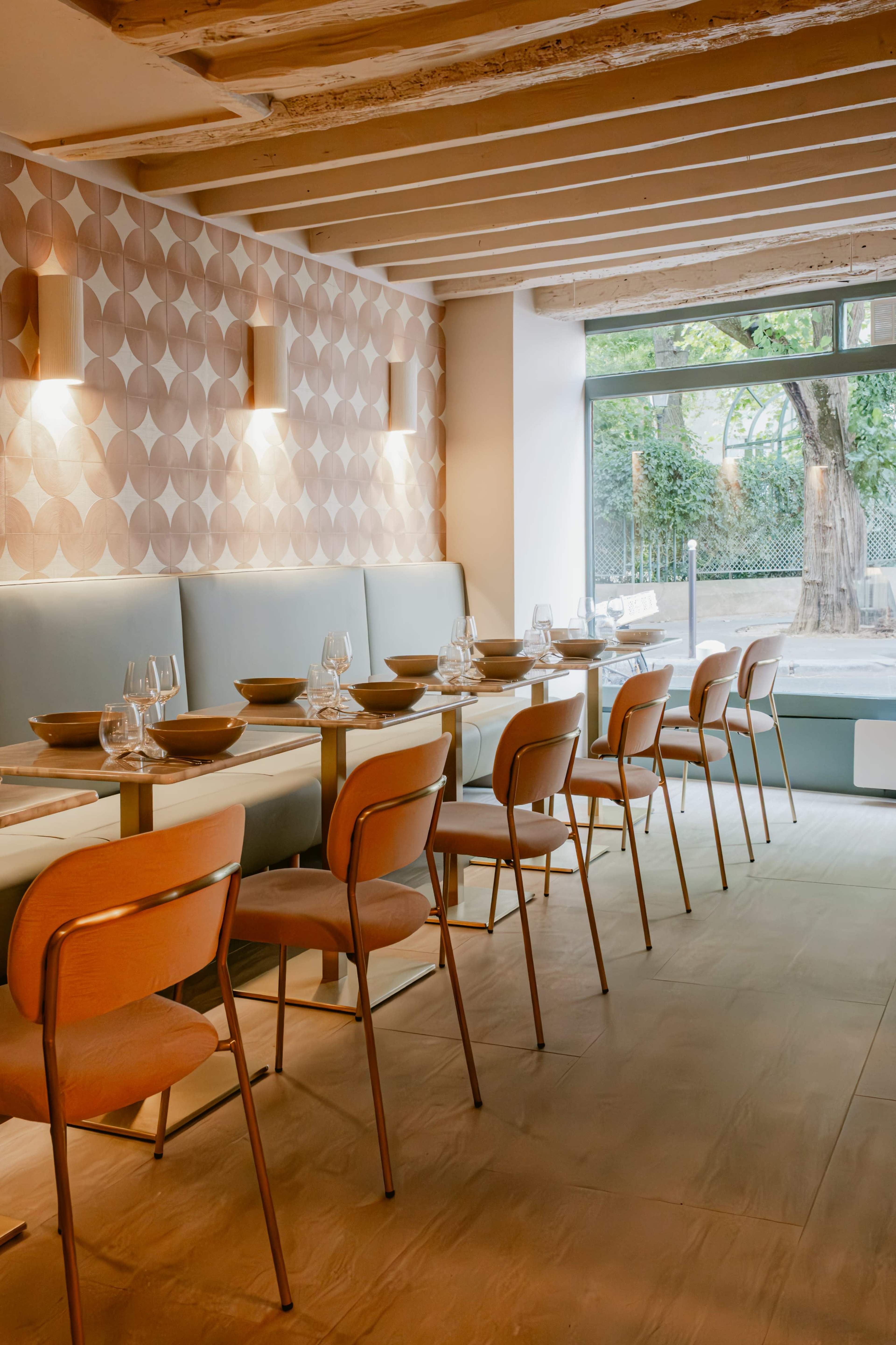 The image shows a modern restaurant interior with a row of elegantly set tables and minimalist decor, featuring a patterned wall and large windows that allow natural light to enter.
