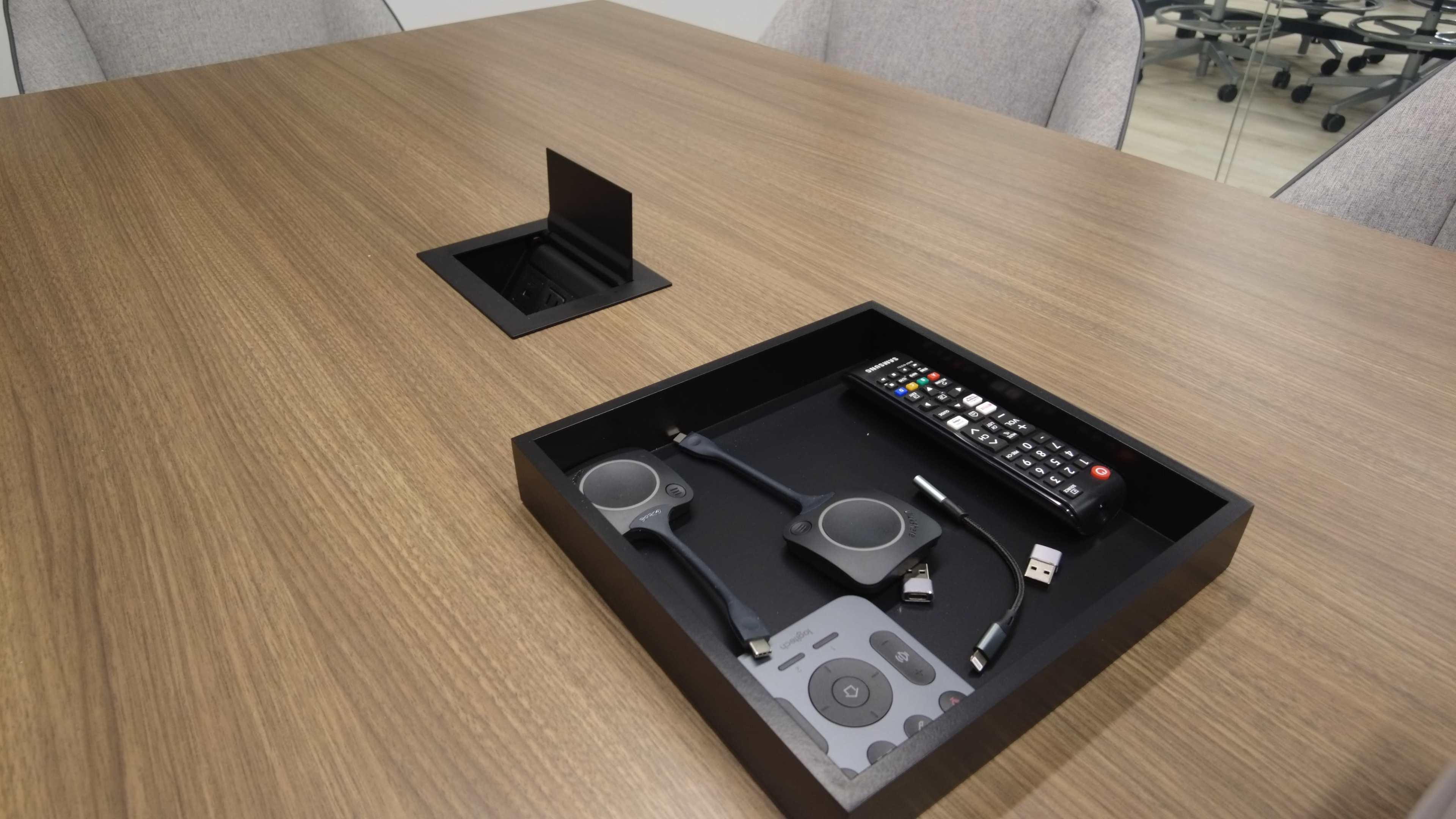 A conference table features a recessed box containing a remote control and various electronic cables.