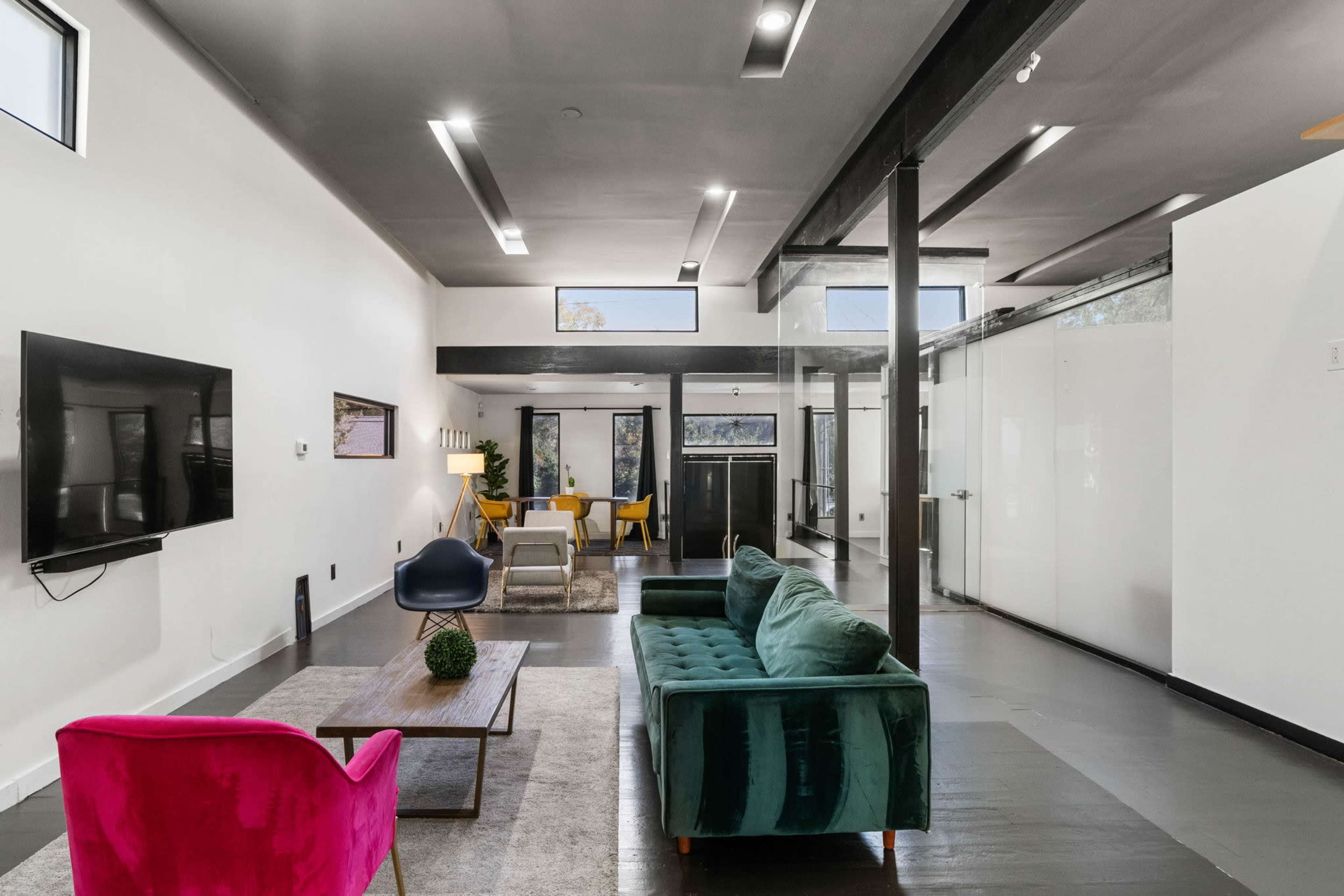 The image shows a modern living space with a green velvet sofa, a pink accent chair, a coffee table, and a television mounted on the wall, all arranged in an area with large windows and an open design.