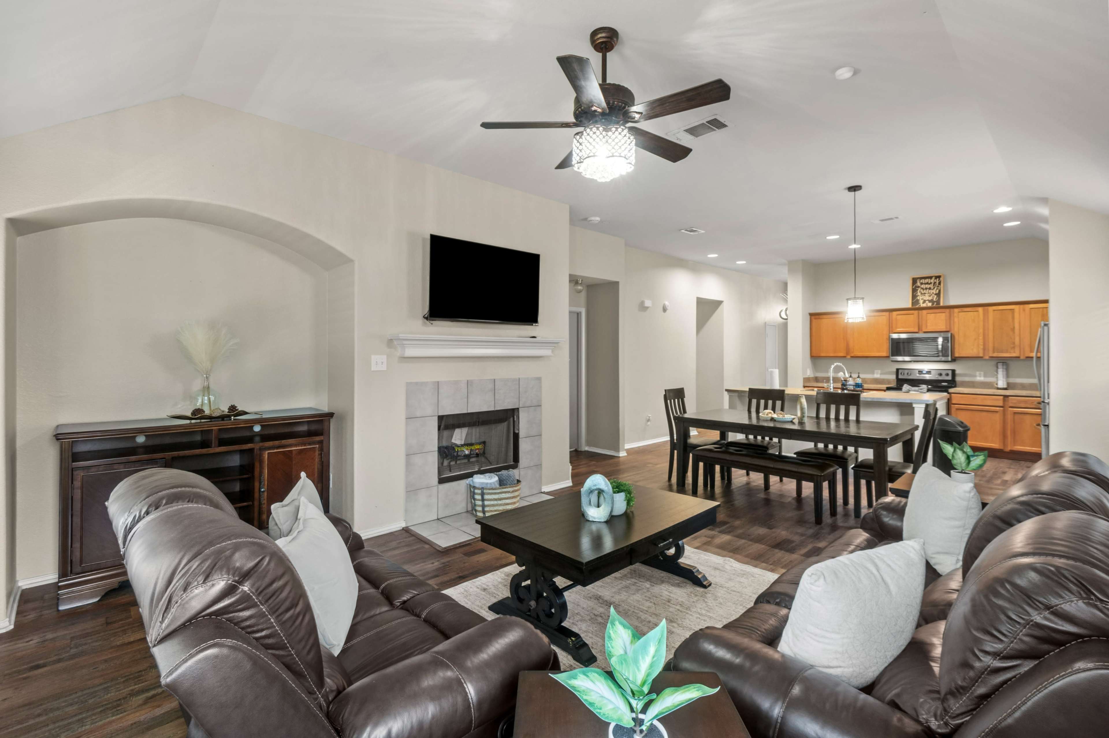 The image shows a cozy living room with leather couches, a coffee table, and a television mounted above a fireplace, leading into a dining area and kitchen with wooden cabinets.