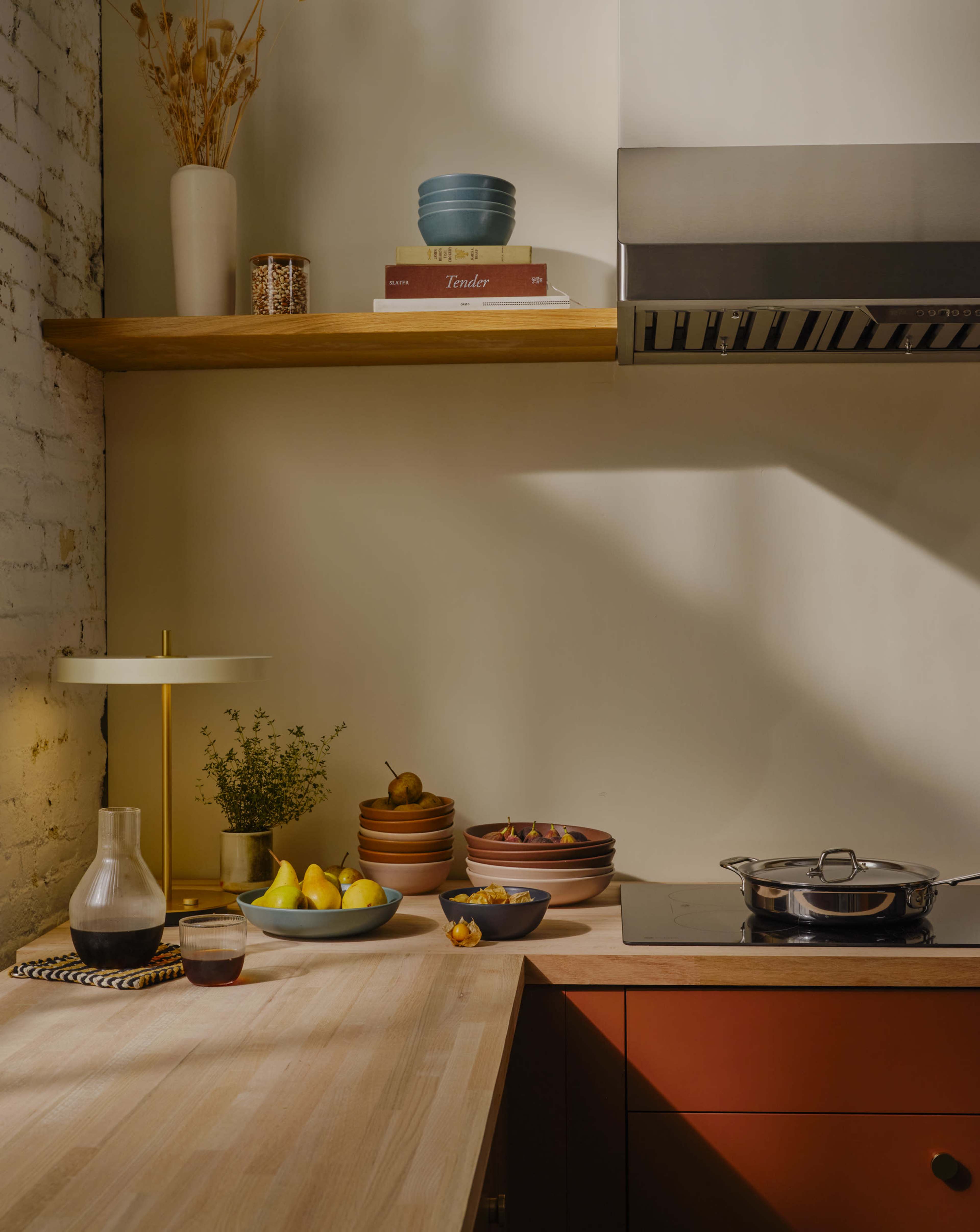 A well-organized kitchen scene features a wooden countertop with various plates of fruit, a cooking pot on the stove, and decorative items on a shelf above.