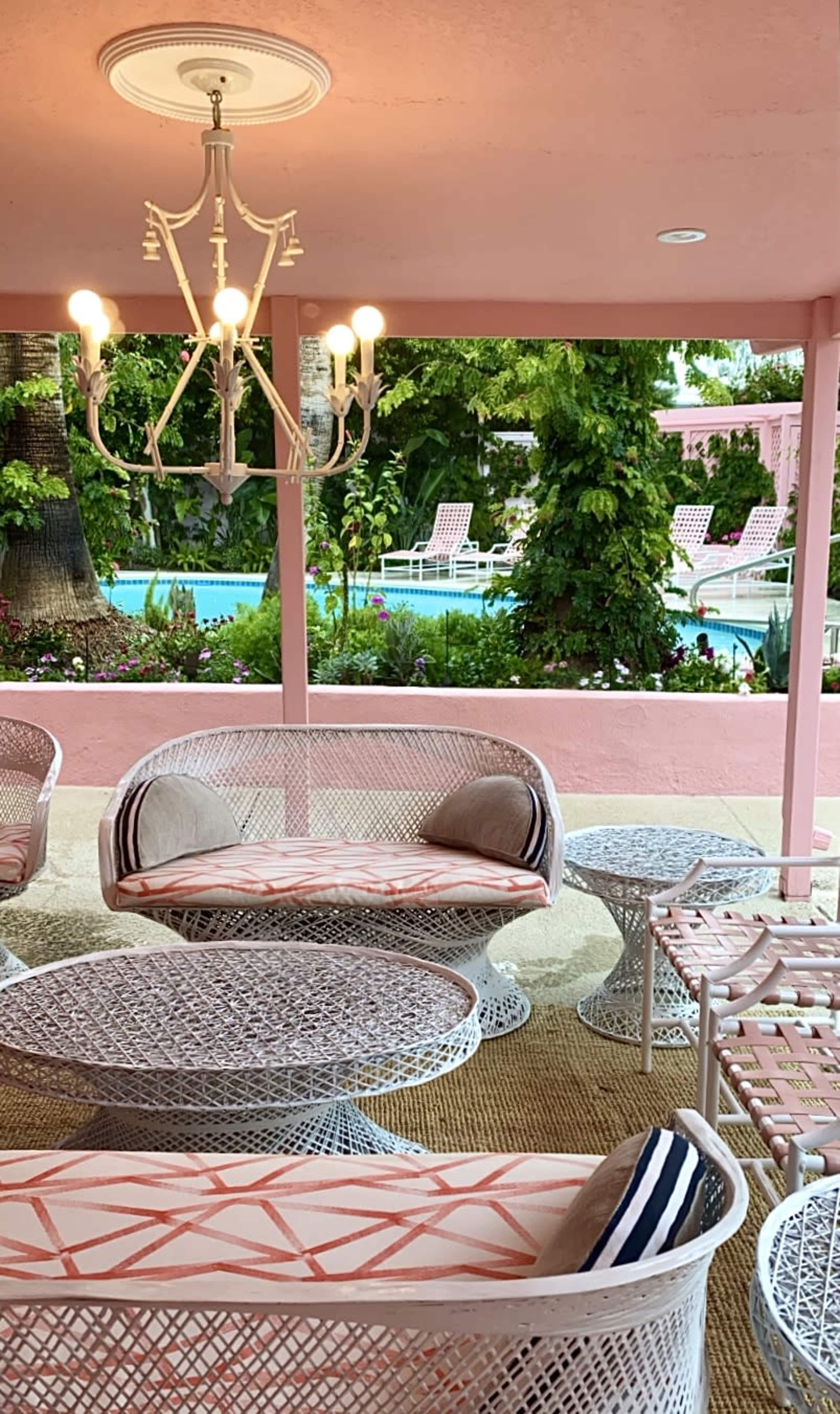 A patio area features white wicker furniture and a chandelier overlooking a pool surrounded by greenery.