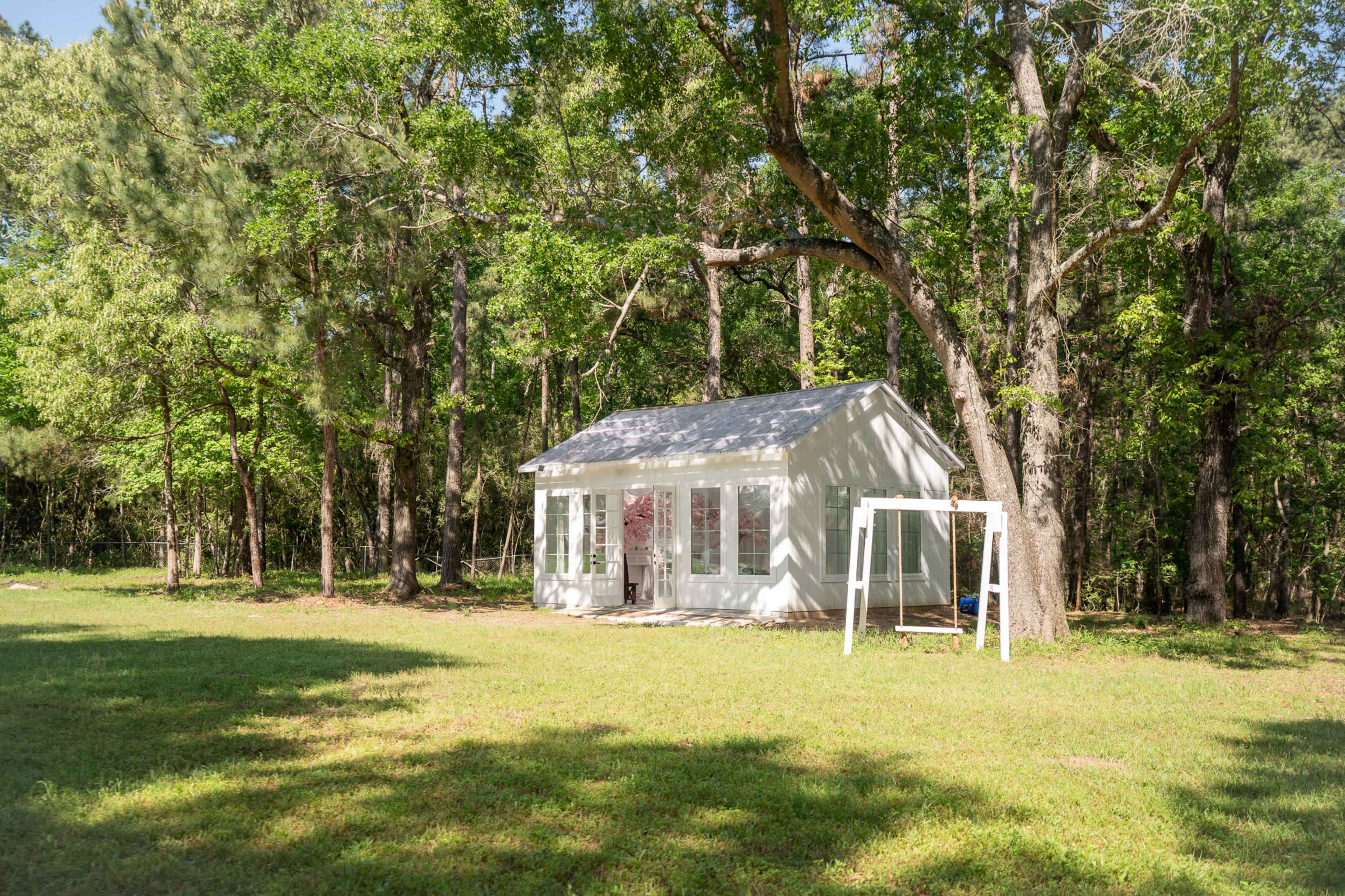 A small white cottage with large windows stands in a grassy area surrounded by trees.